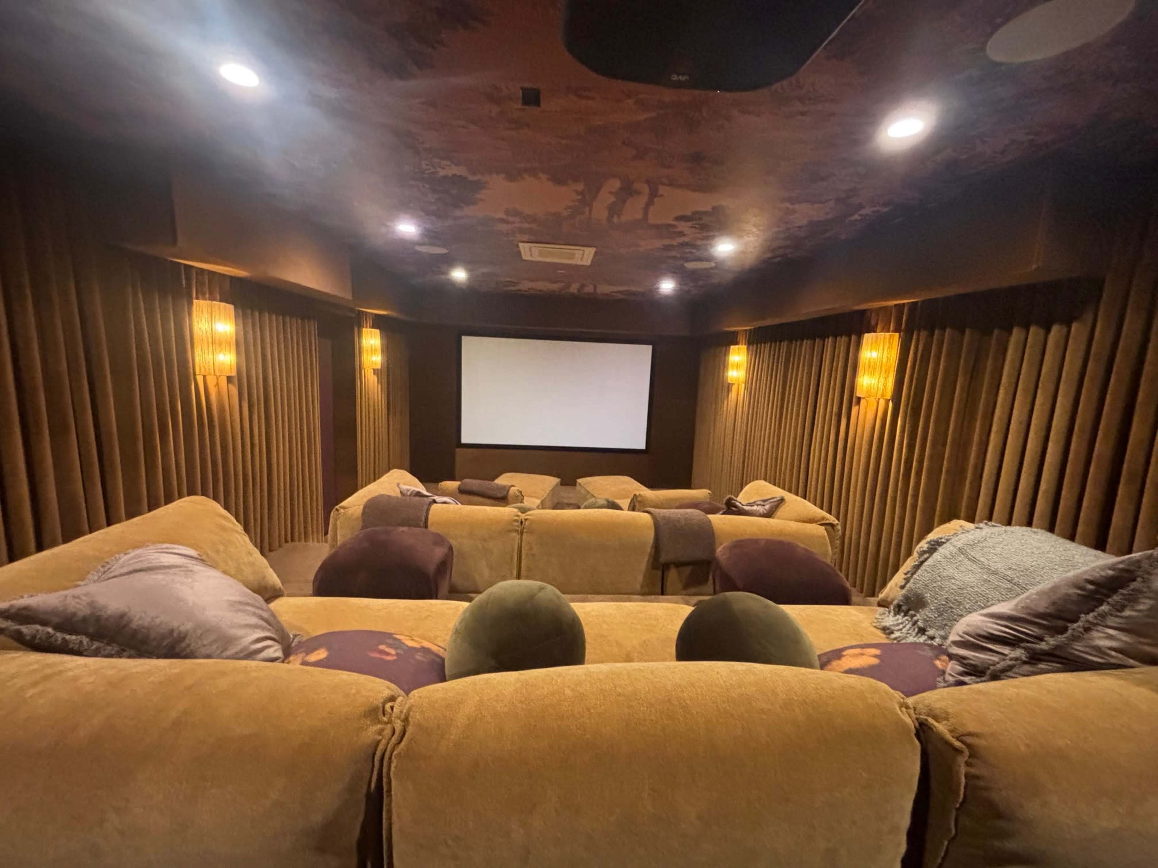 The image shows a cozy home theater with plush seating arranged in front of a large screen, surrounded by curtained walls and ambient lighting.