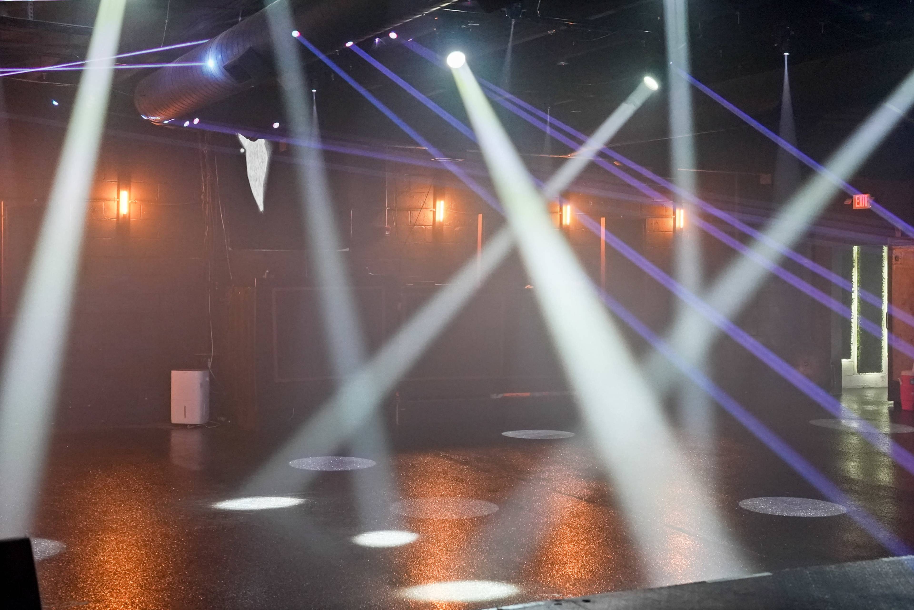 The image shows a dimly lit performance venue with beams of light cutting through the fog, highlighting a polished floor.