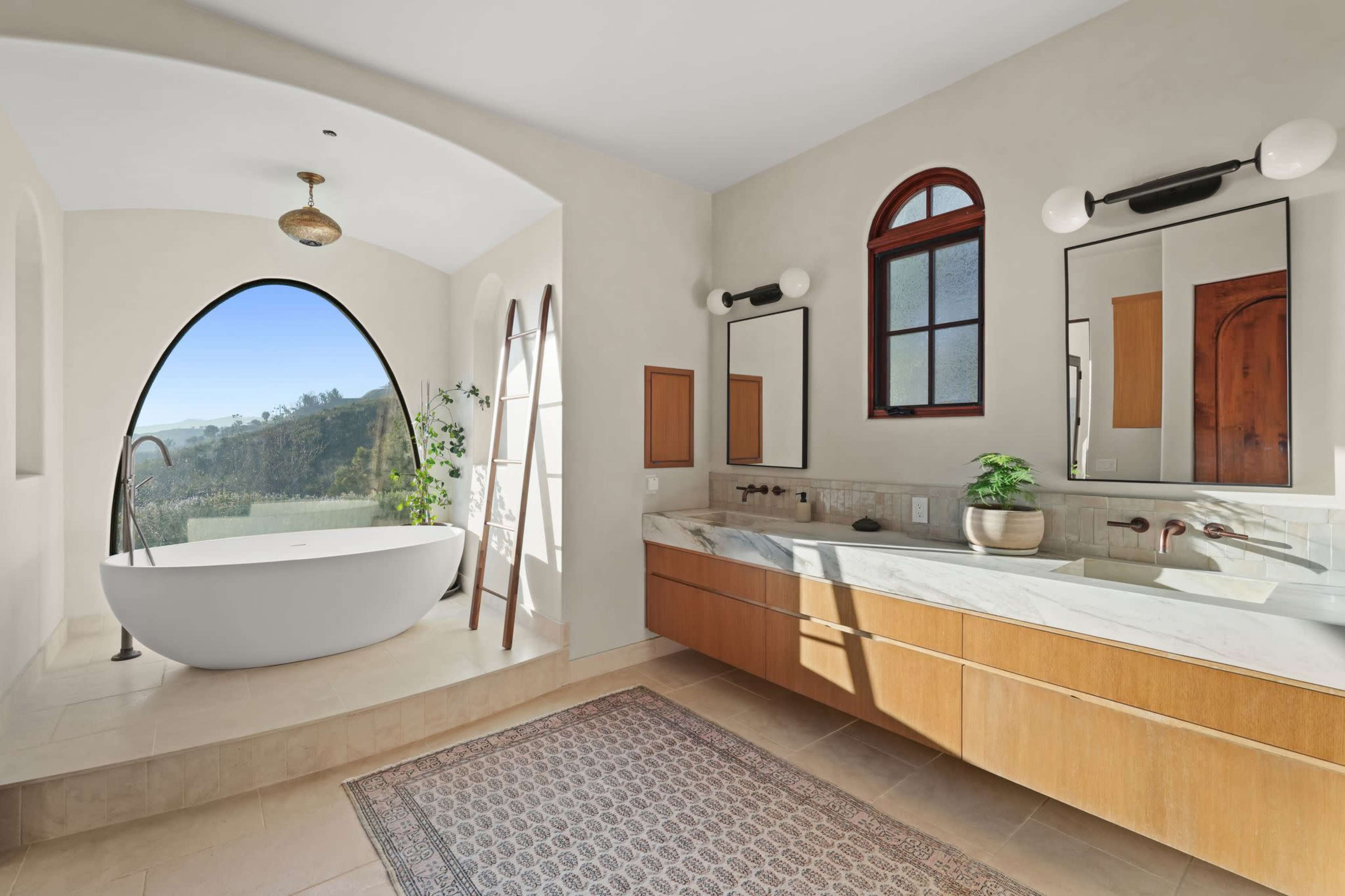A modern bathroom features a freestanding bathtub beside a large arched window, wooden cabinetry, and dual sinks with mirrors.