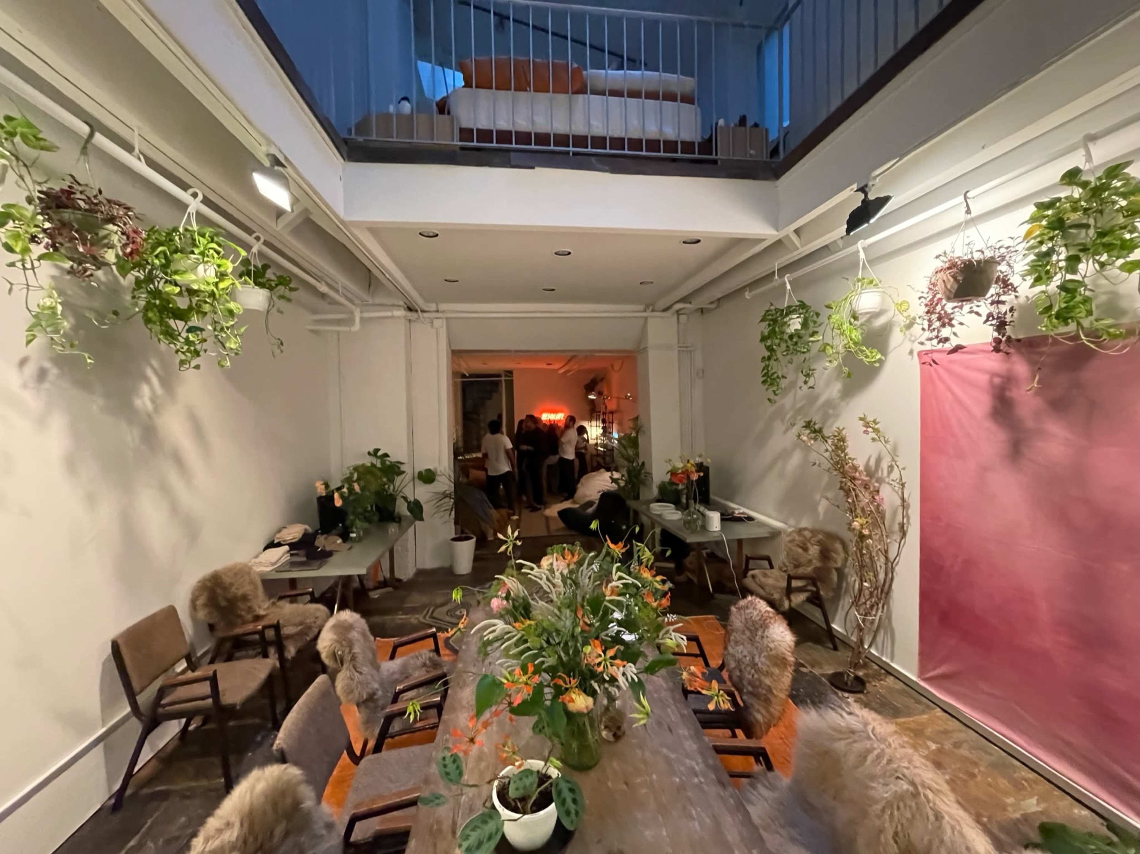 A well-decorated indoor space with a long dining table surrounded by chairs, plants hanging from the ceiling, and a visible upper level with a bed.