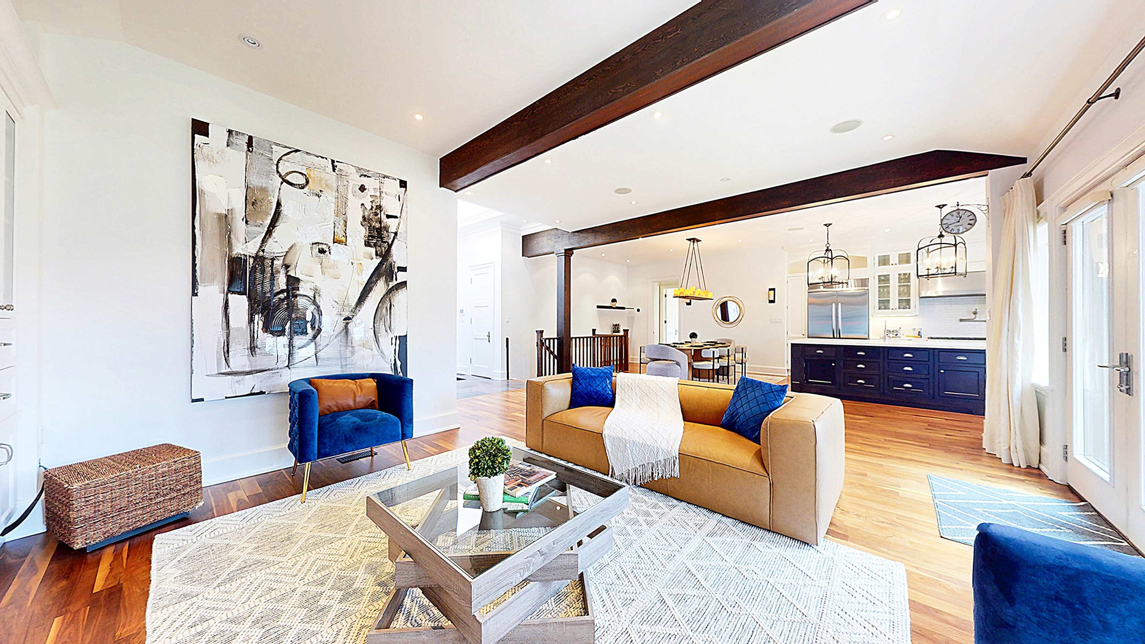 The image shows a modern living room featuring two blue armchairs, a beige sofa, a wooden coffee table, and a large abstract painting on the wall, with a staircase and kitchen visible in the background.