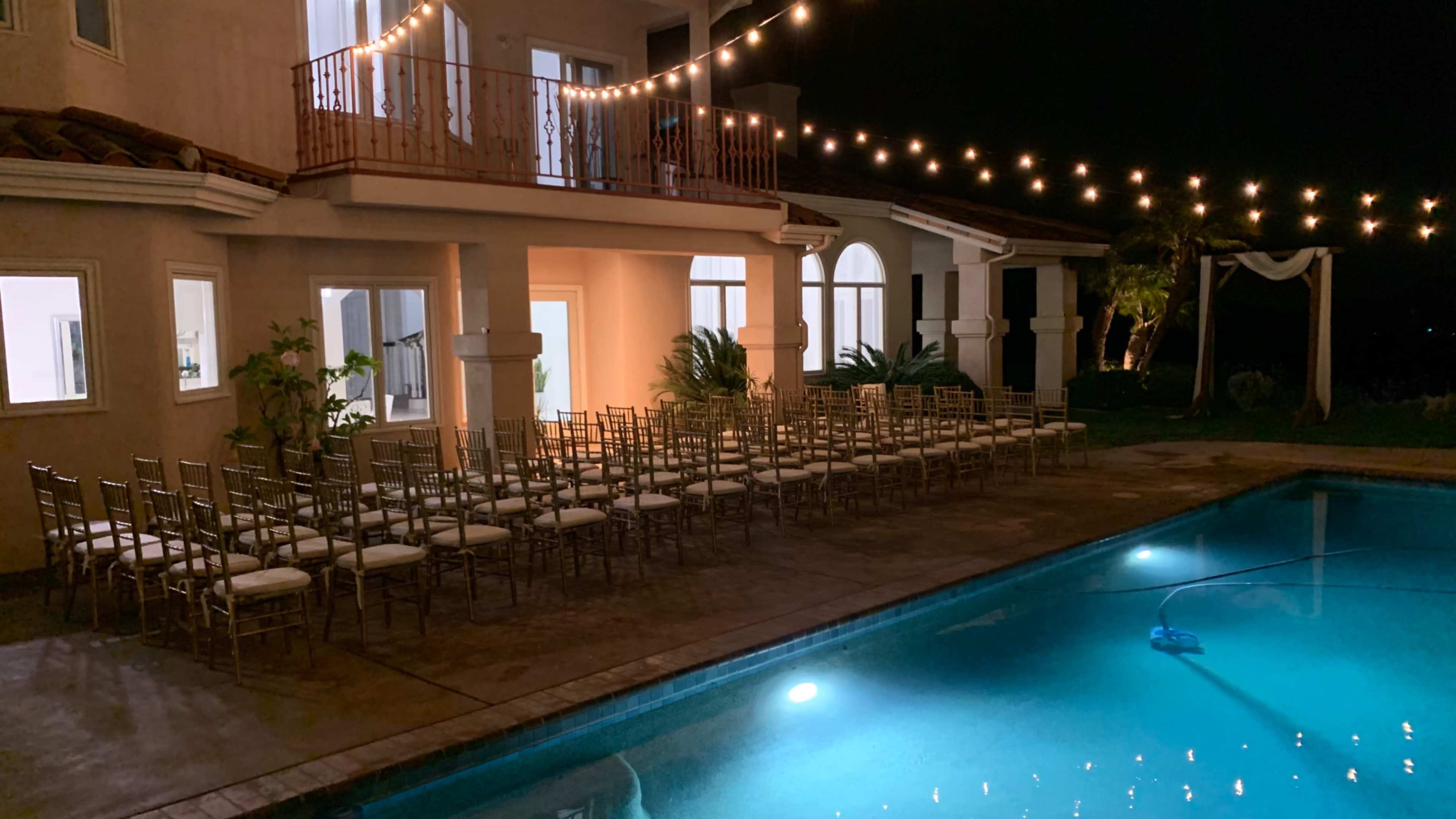 The scene features a well-lit outdoor area with a swimming pool, adjacent to a house, and several rows of empty chairs set up for an event under string lights at night.