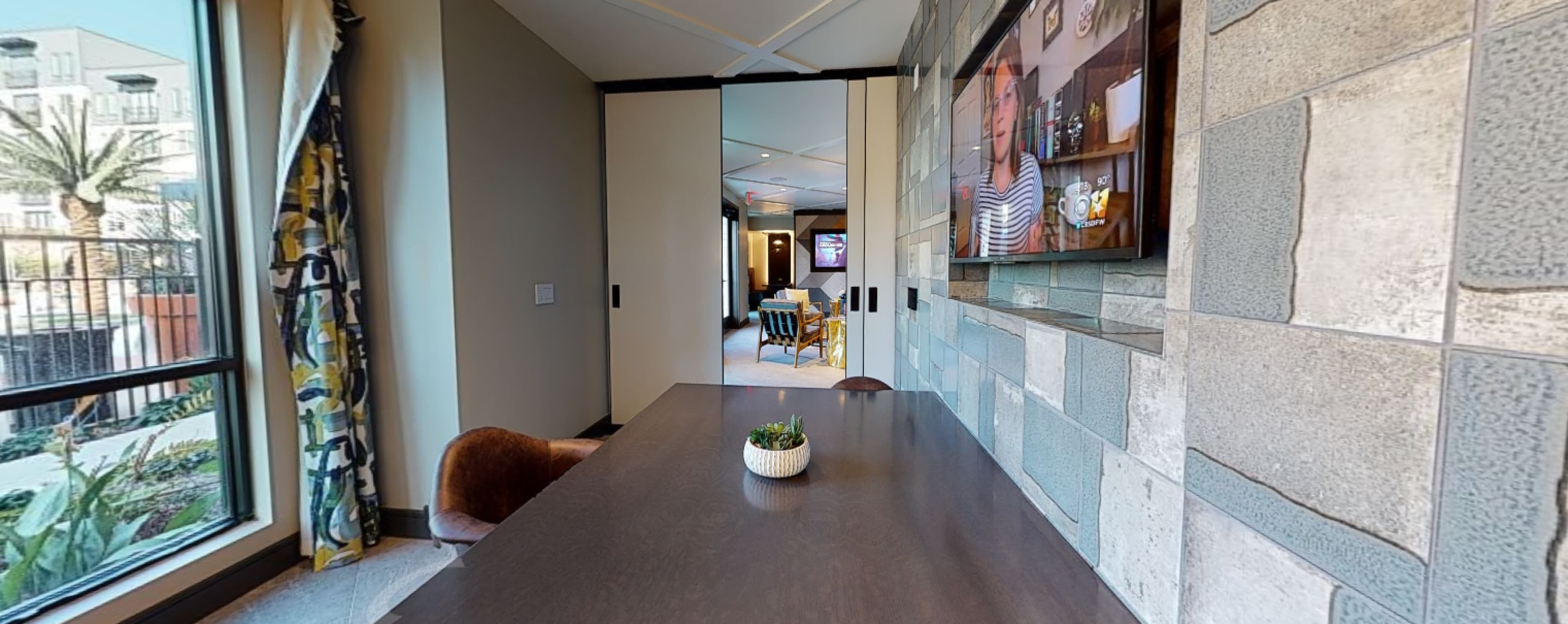 The image shows a modern interior space featuring a table with a small plant, a TV mounted on a wall, and a view of an outdoor area through large windows.