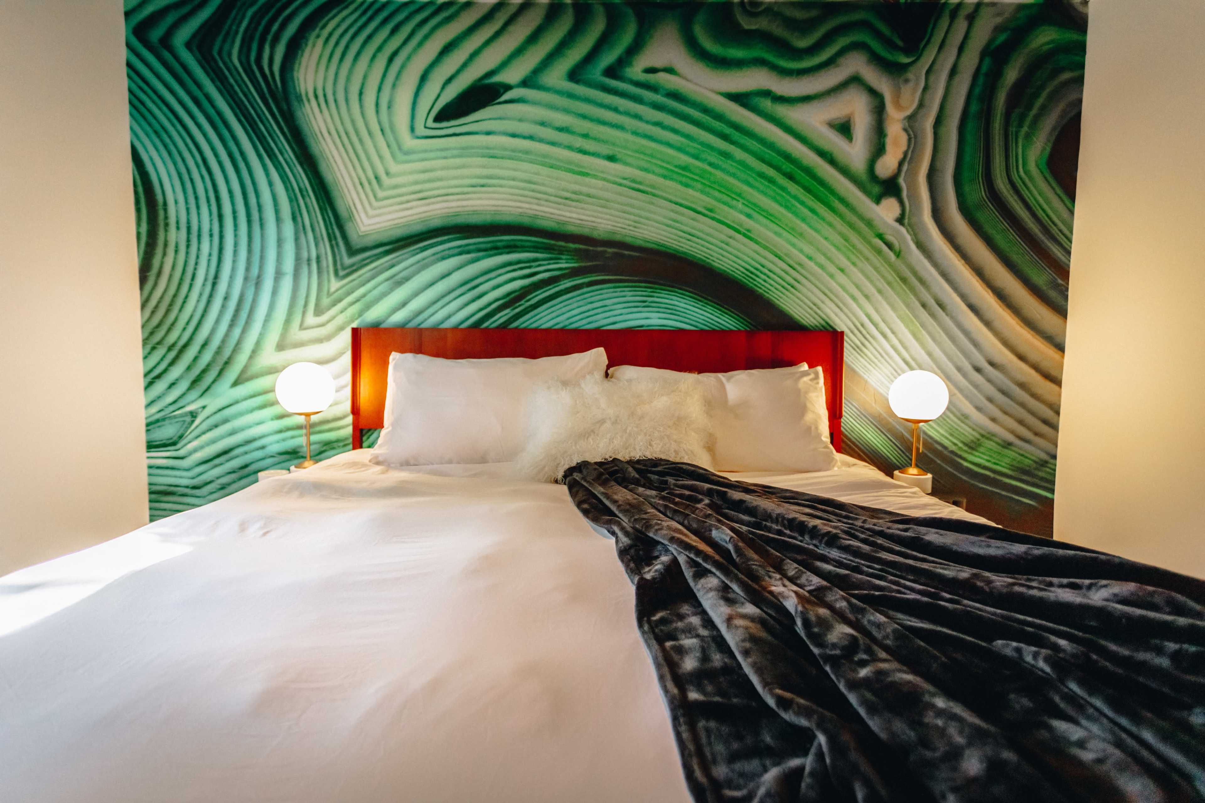 A neatly made bed with a dark blanket, flanked by two bedside lamps, against a textured green wall.