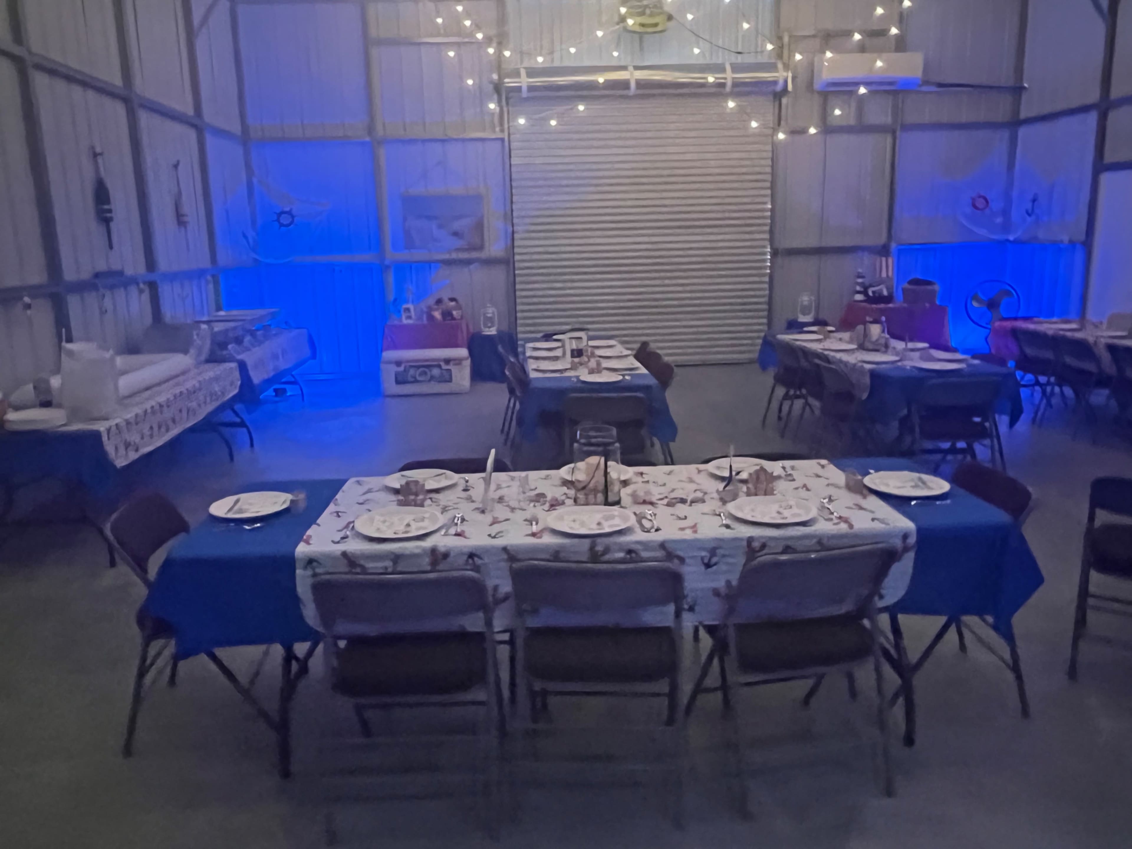 The image shows a spacious indoor dining area with several tables covered in blue tablecloths, set for a meal, and illuminated by soft blue lights.