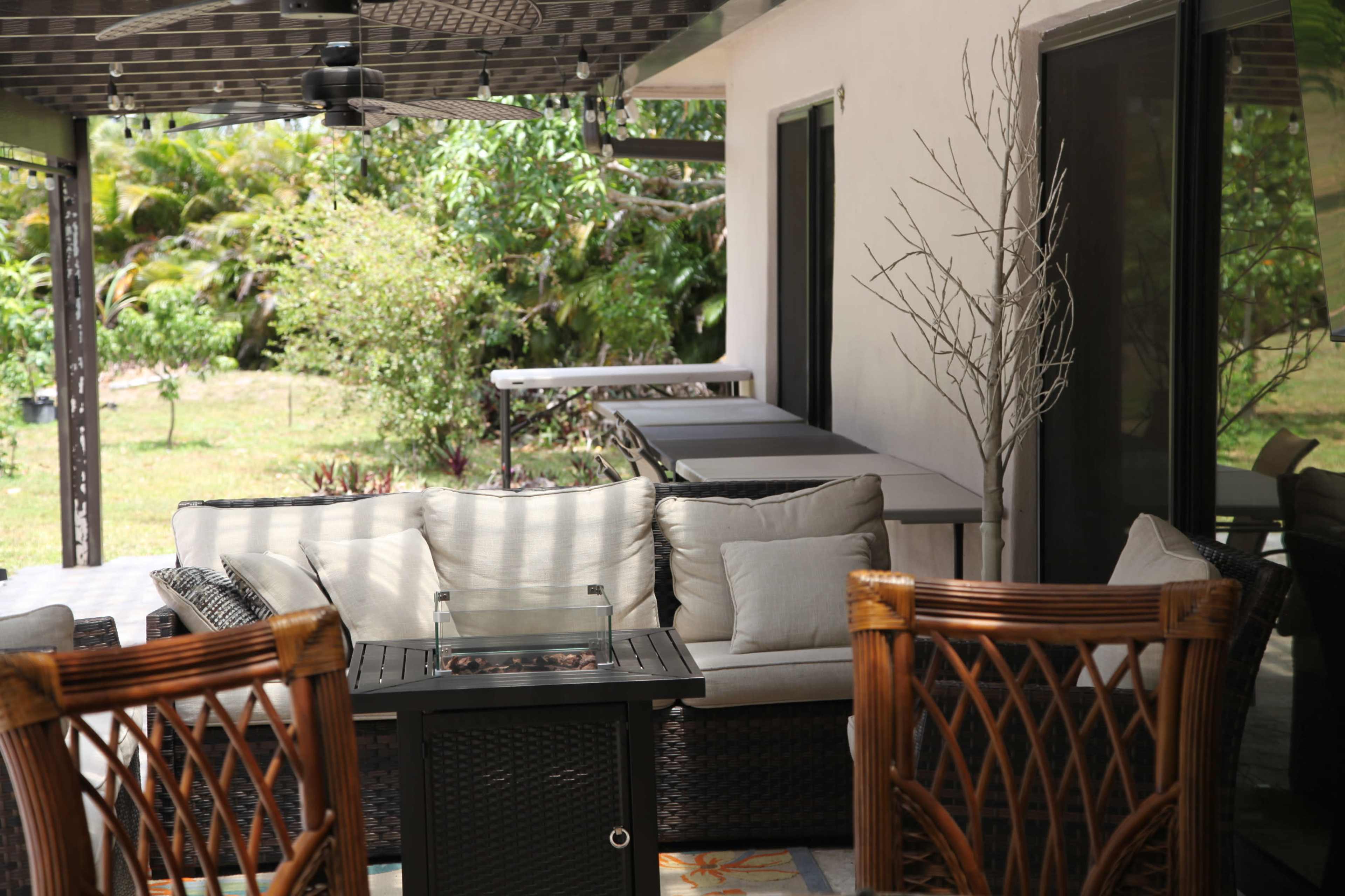 A patio area with a woven seating arrangement, a fire table, and a view of a grassy backyard surrounded by greenery.