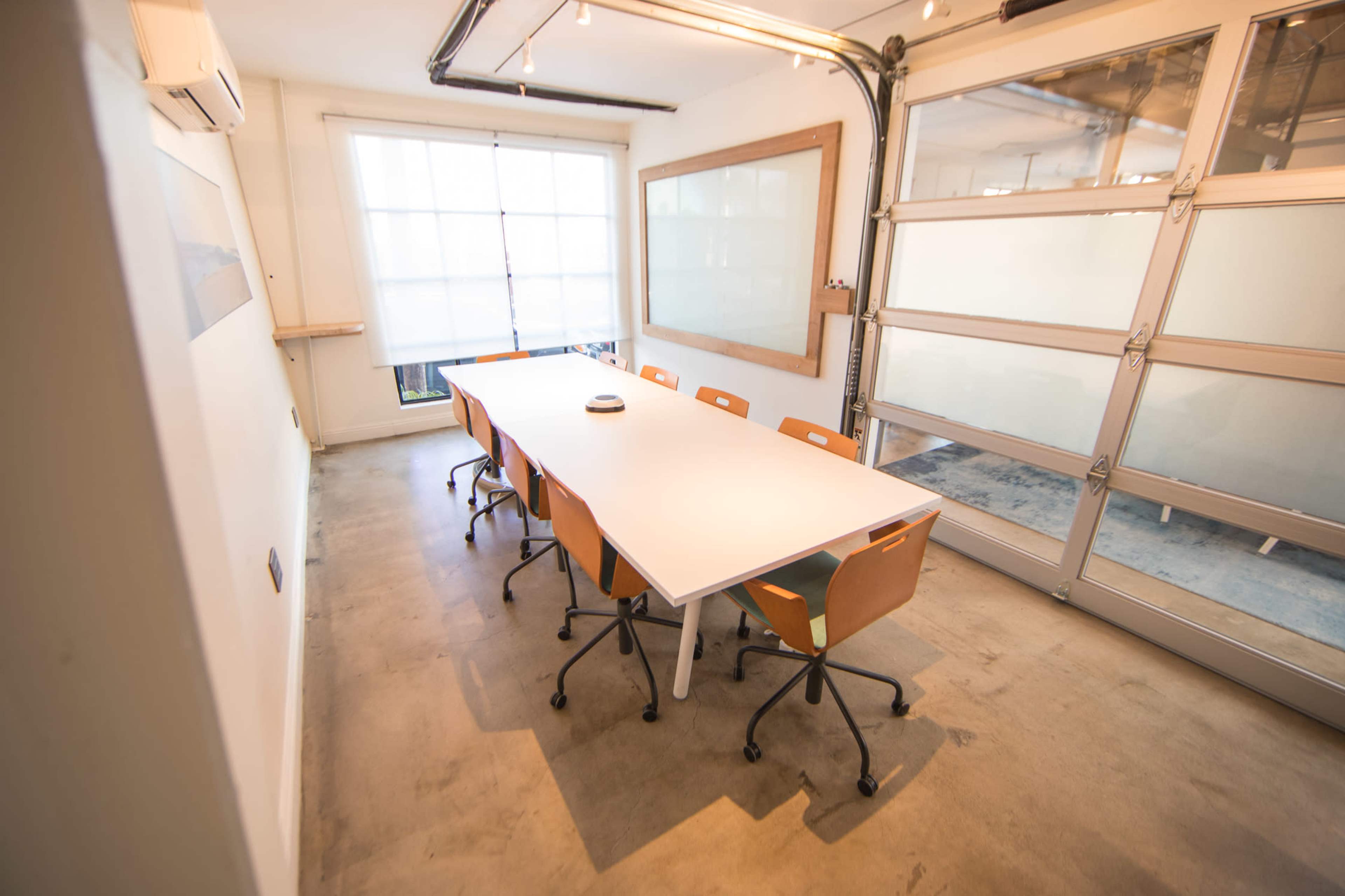 A modern conference room with a long white table surrounded by several orange chairs, large windows, and a glass garage door.