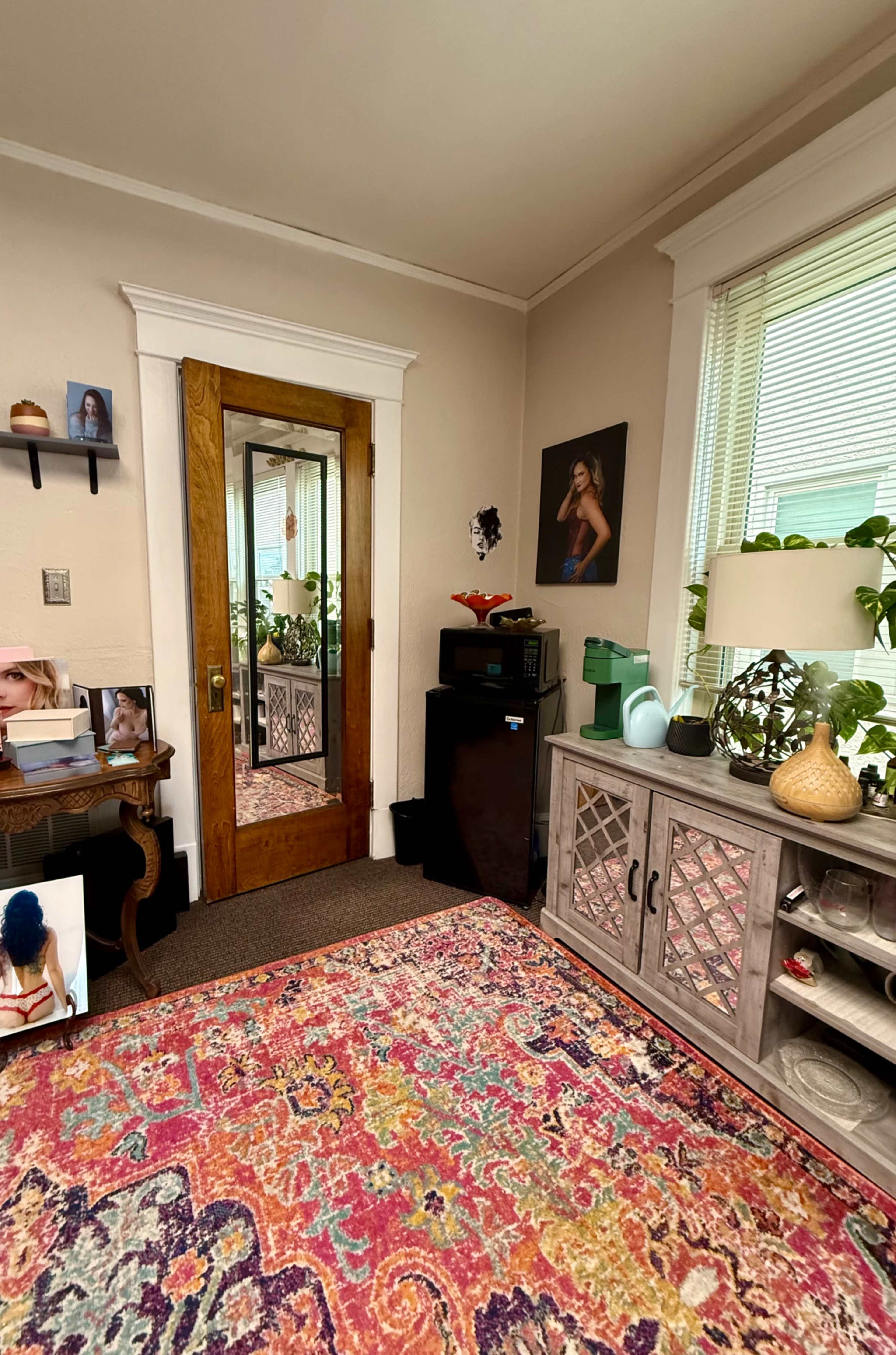 The image shows a cozy room featuring a wooden door, a mirror, a decorative cabinet, a lamp, plants, and colorful artwork on the walls.