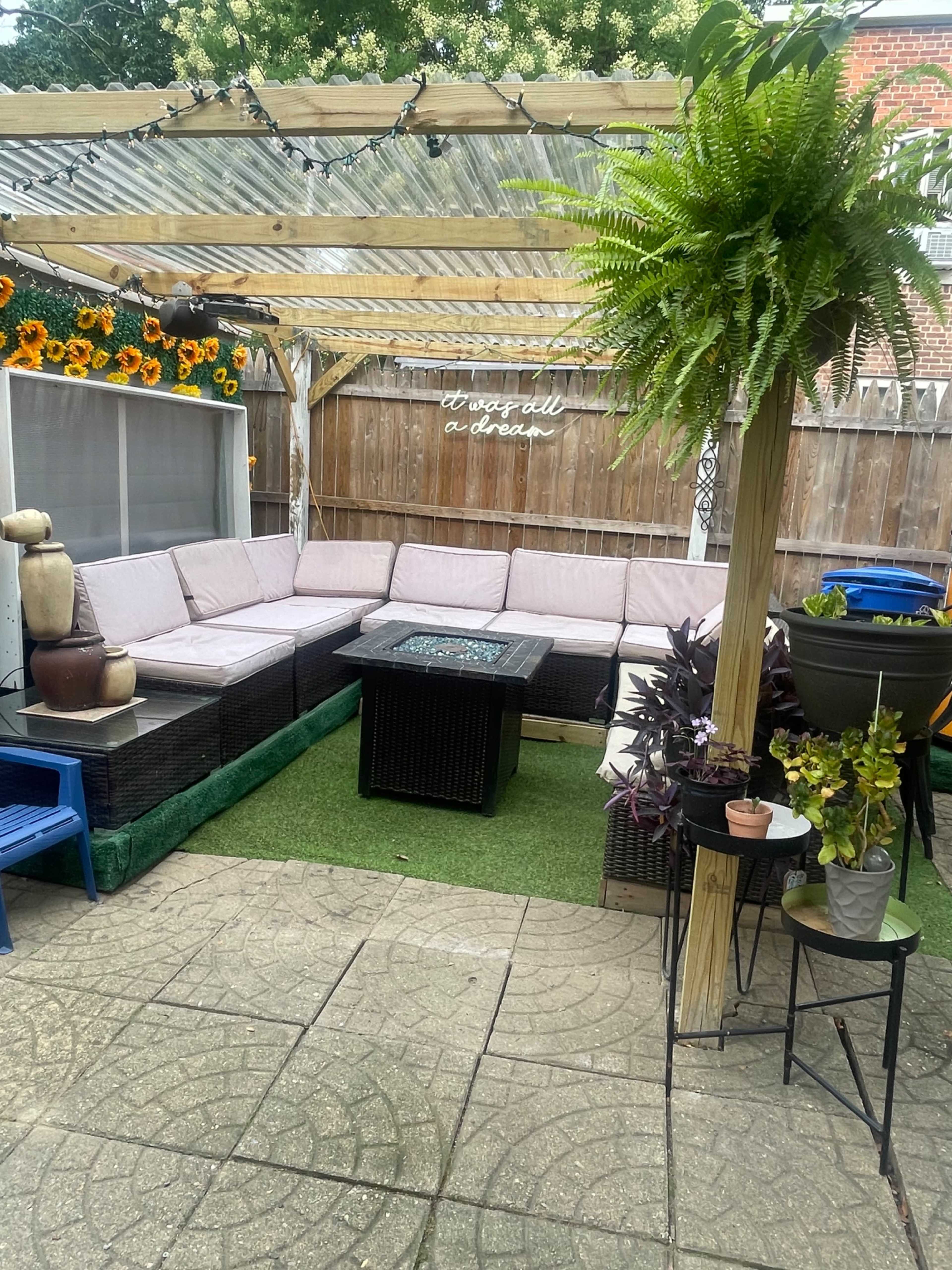 A patio area with a large sectional sofa, a coffee table in the center, and various potted plants, all under a wooden pergola adorned with string lights.