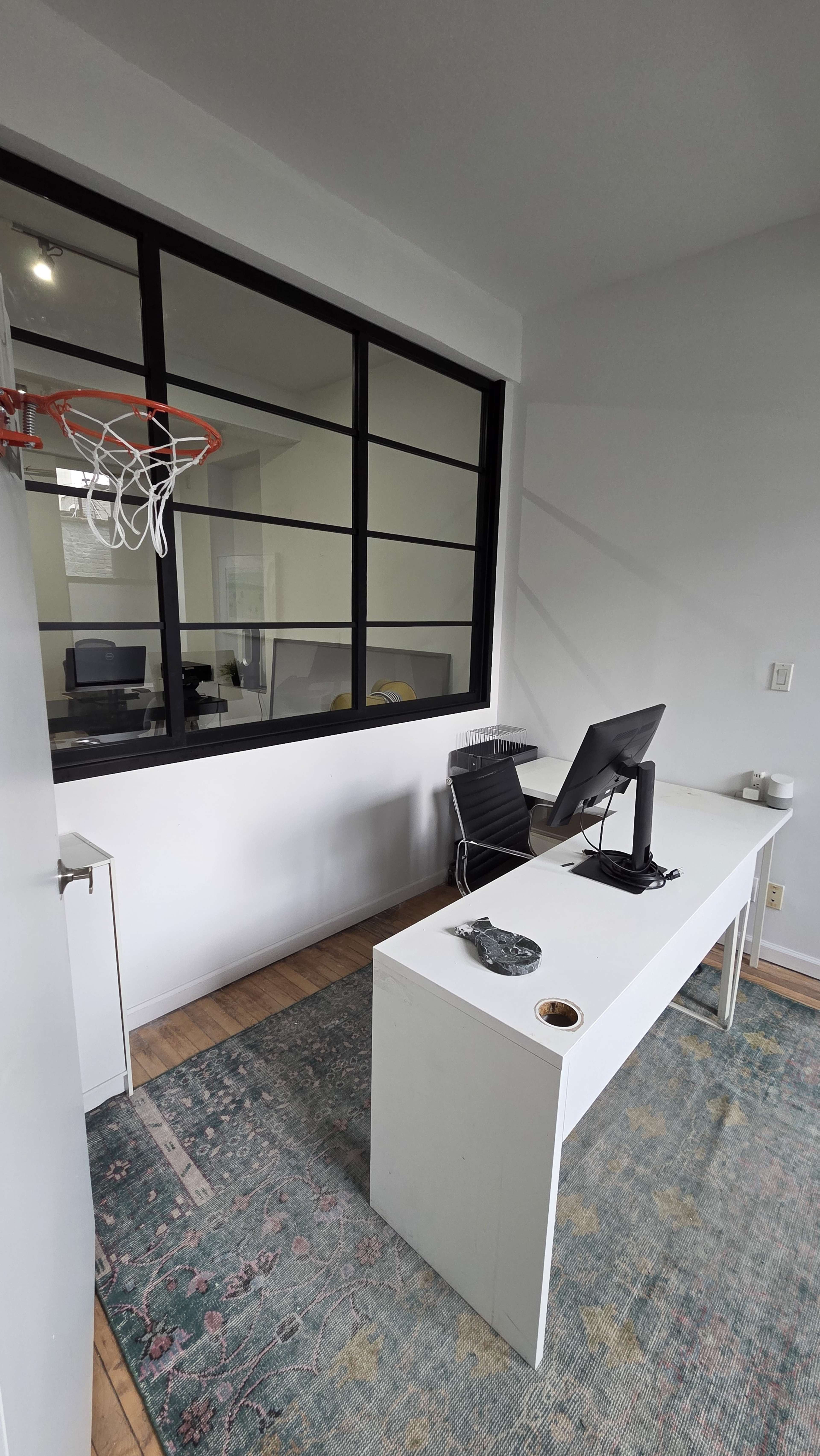 The image depicts a modern office space featuring a desk with a computer, a black chair, and a basketball hoop mounted on the wall.