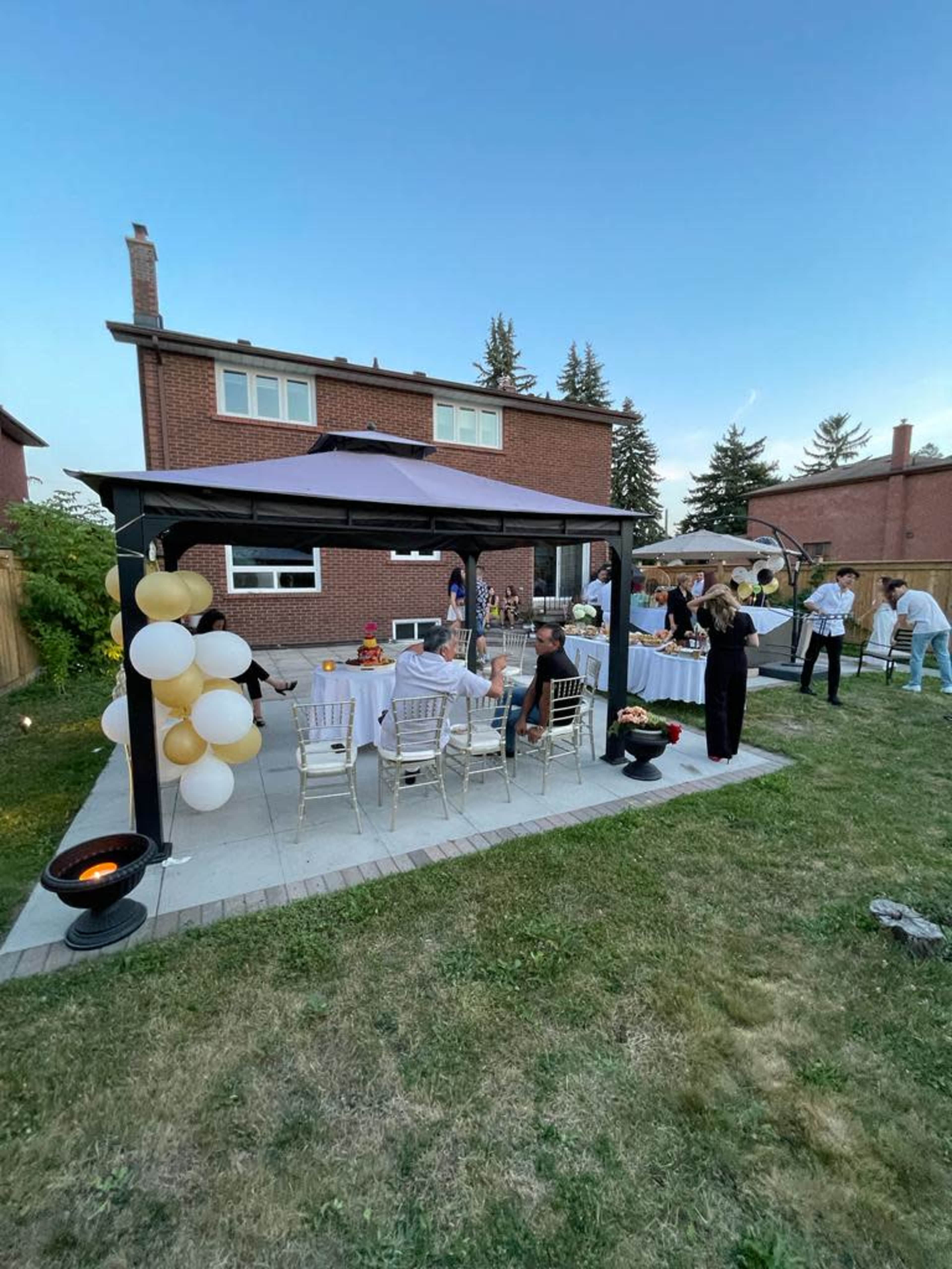 A group of people is gathered under a gazebo in a backyard, with a table set for an outdoor event decorated with balloons.
