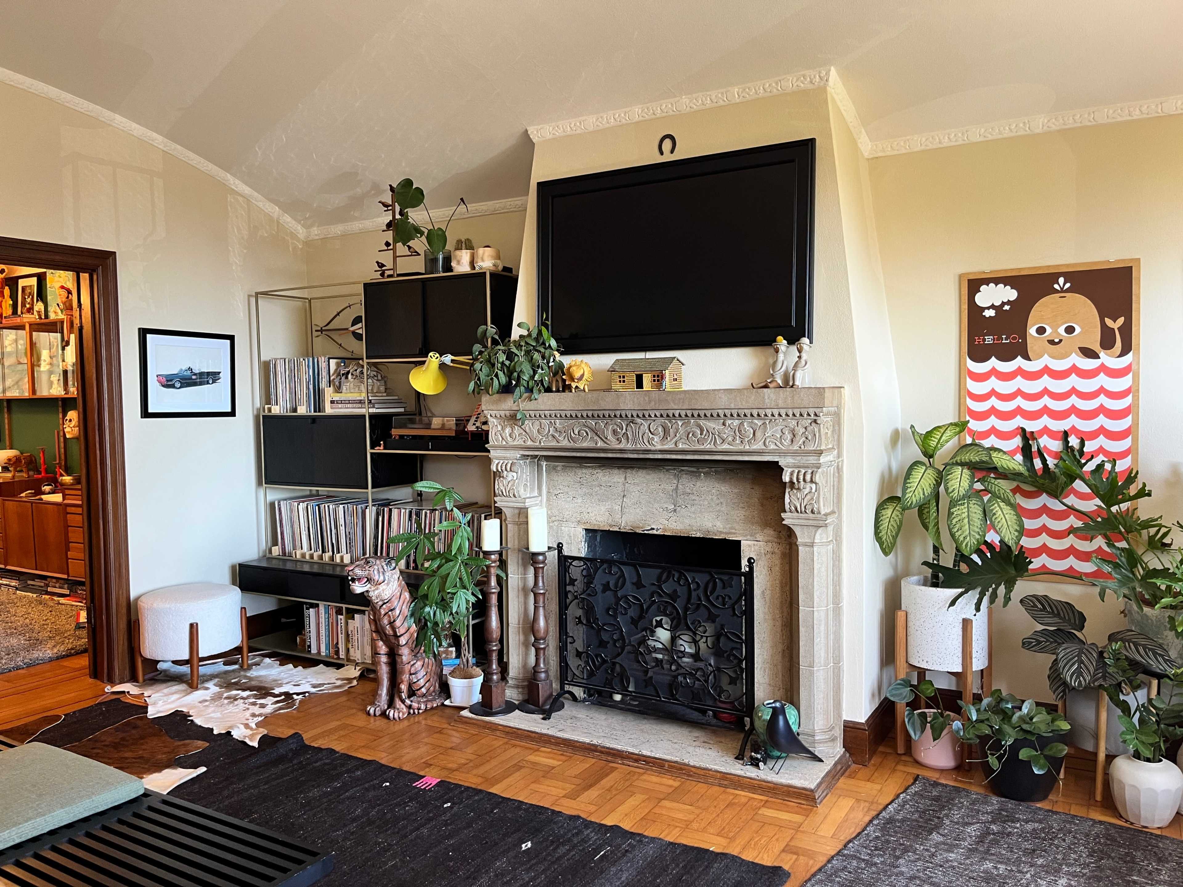 The room features a stone fireplace with a TV mounted above it, surrounded by plants, bookshelves, and a decorative wall with artwork.