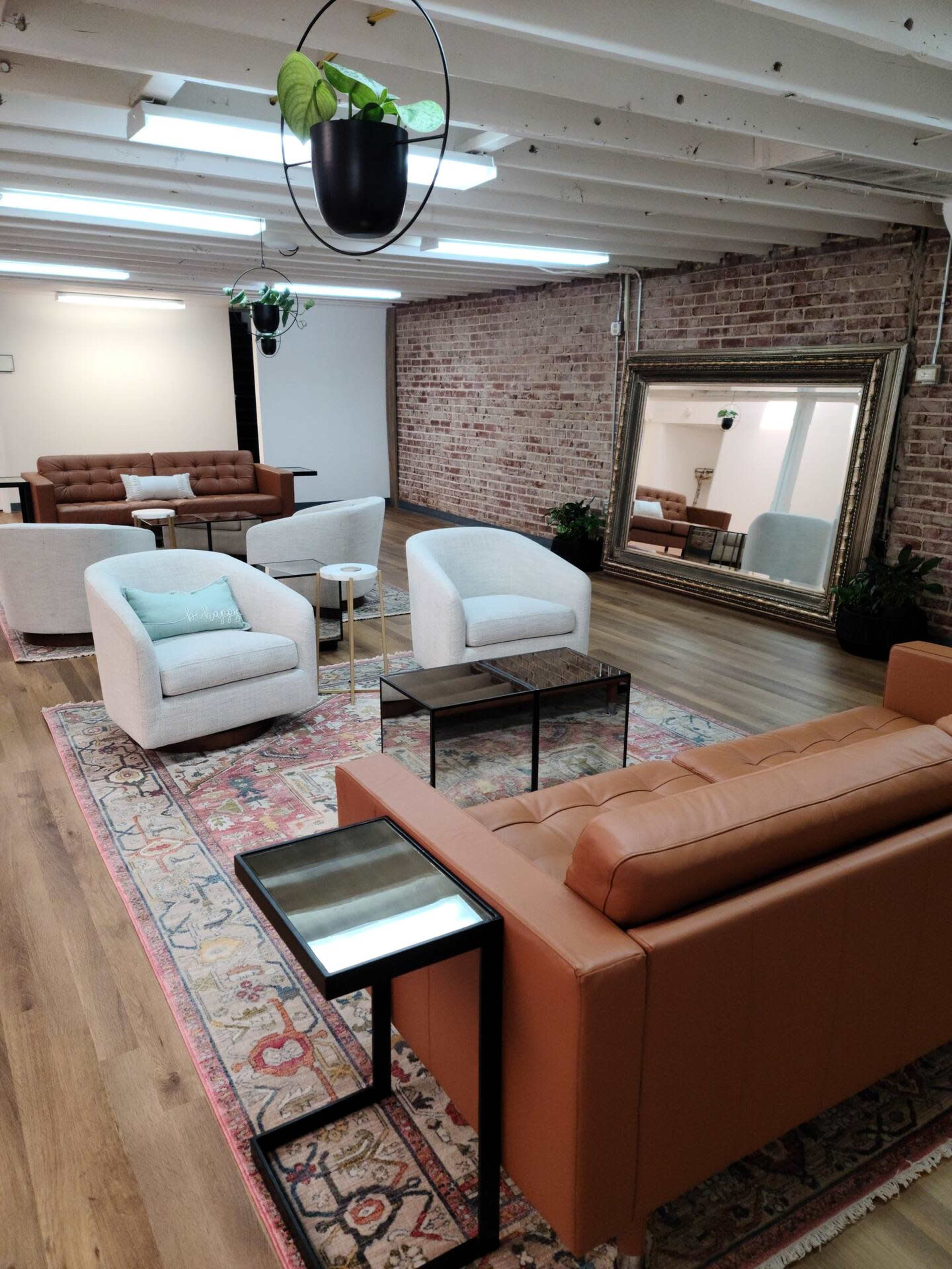 A spacious lounge area with brick walls, featuring multiple seating arrangements including a brown leather couch, white armchairs, and a large mirror.