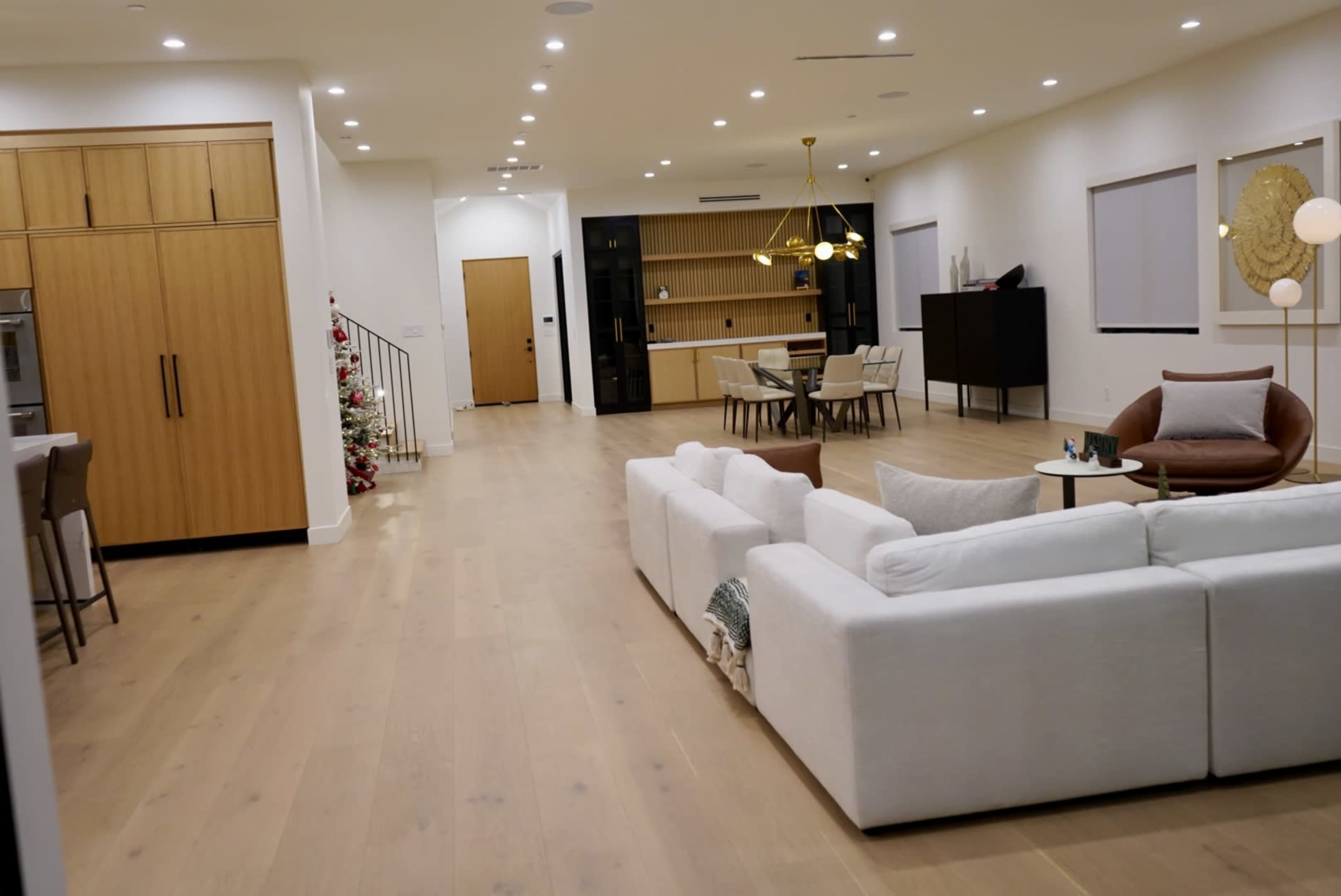 The image shows a spacious, modern living area featuring a white sectional sofa, a brown armchair, and a dining area with shelves and a light fixture.