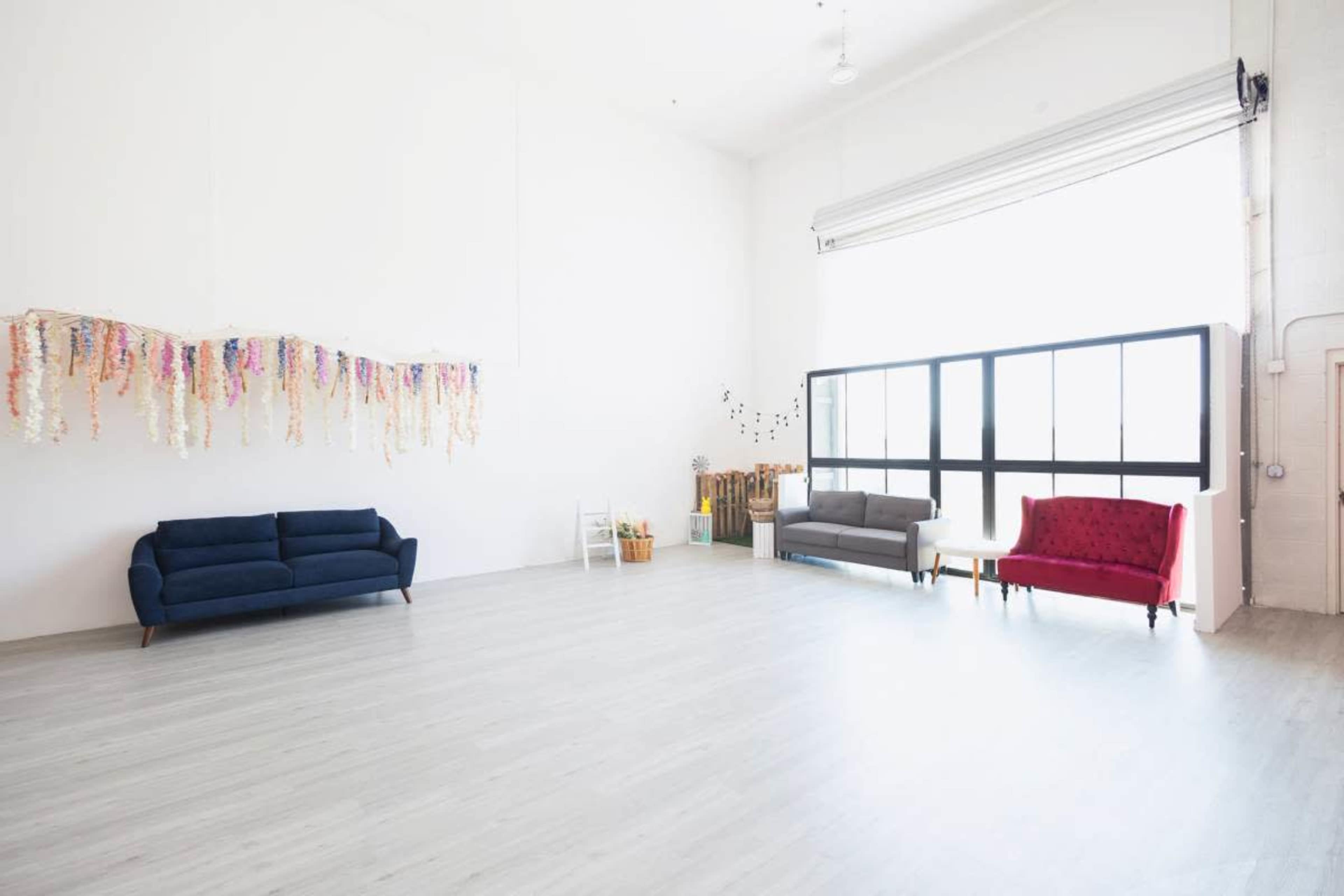 The image shows a minimalist room with light wooden flooring, featuring three couches in varying colors and a decorative wall hanging made of colorful fabric strips.