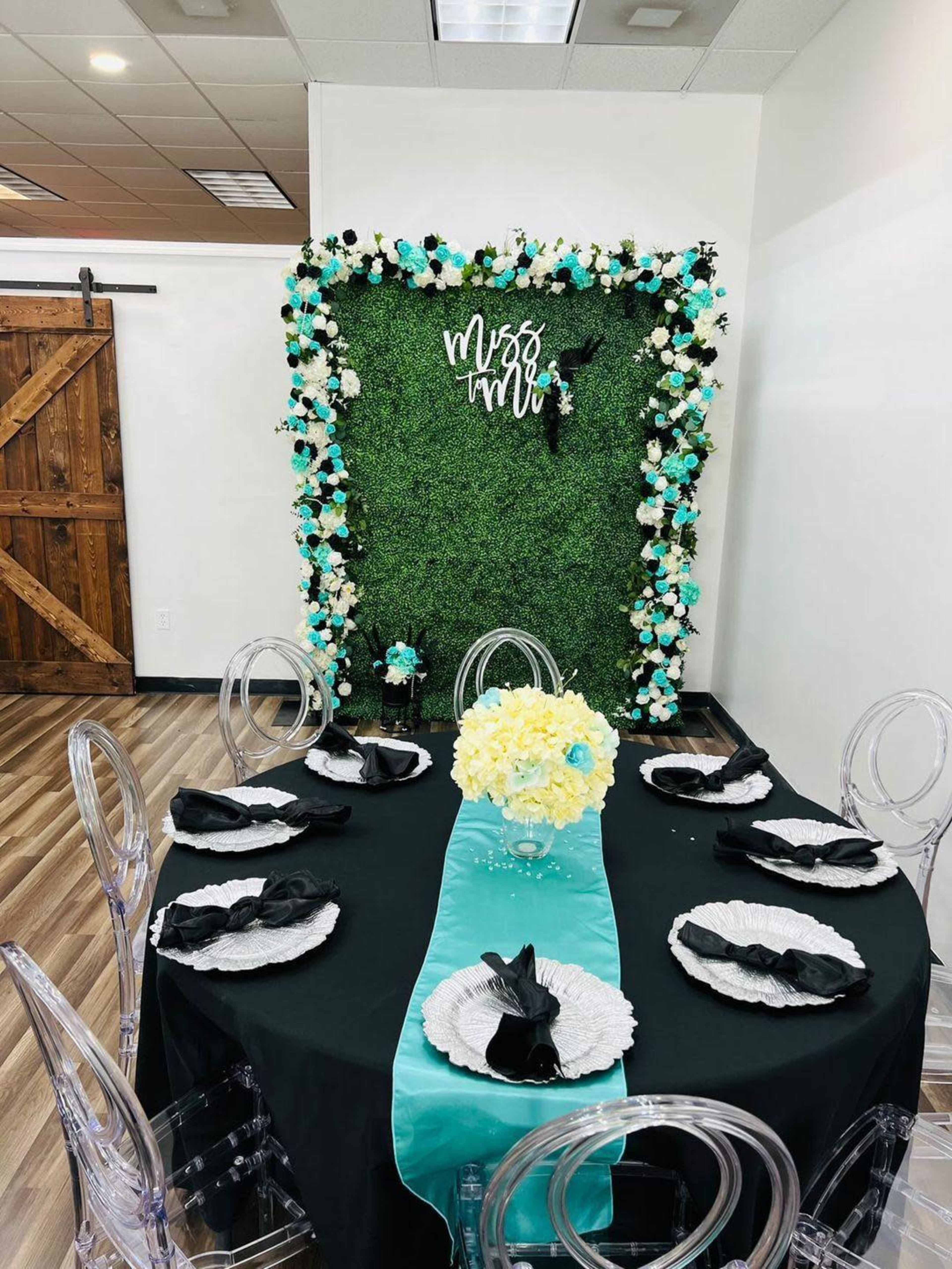 A round dining table is set with black plates and napkins, surrounded by transparent chairs, facing a wall adorned with a floral backdrop that reads "Miss to Mrs."