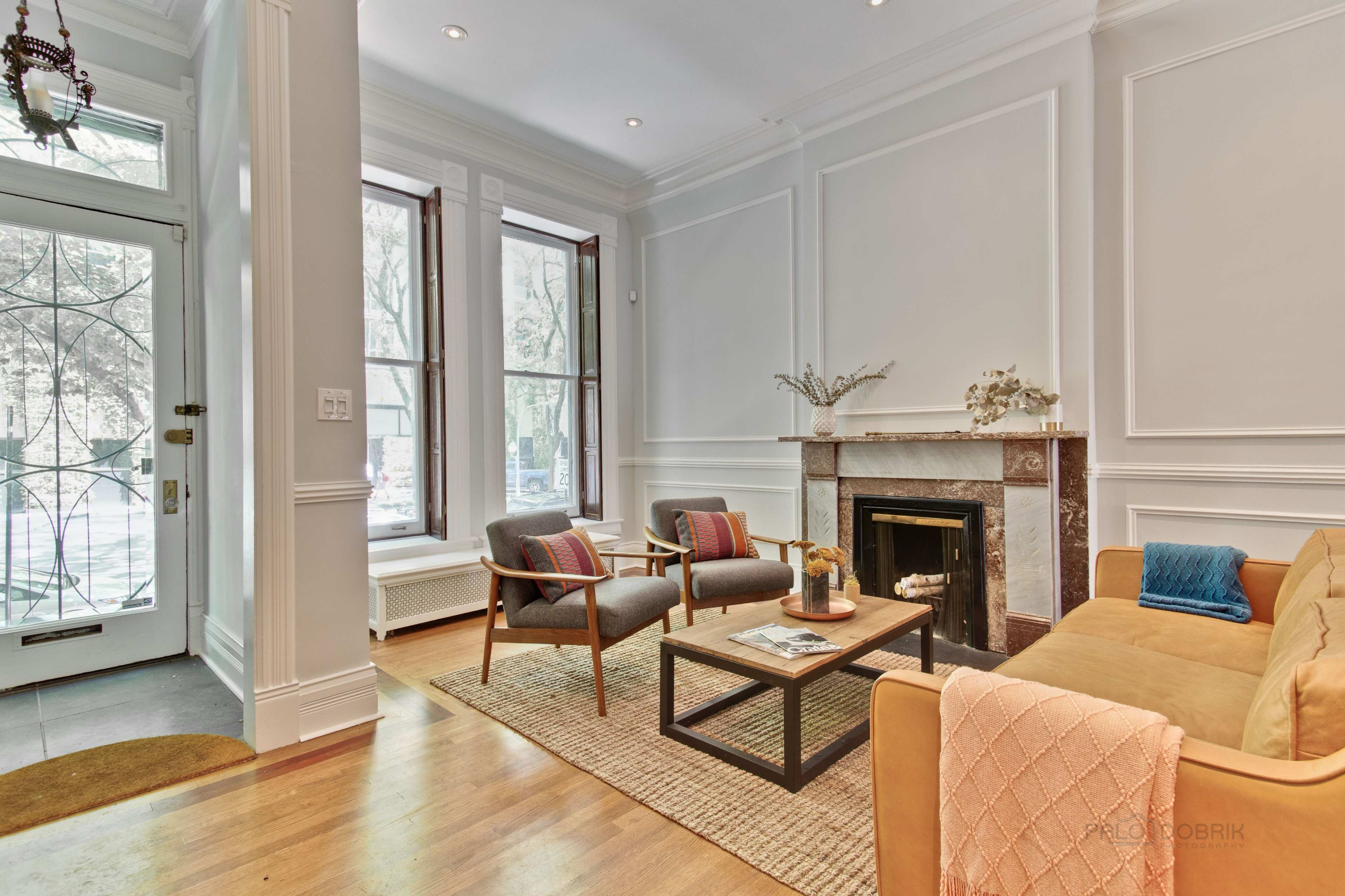 A well-lit living room with a fireplace, two armchairs, a coffee table, and large windows overlooking a street.