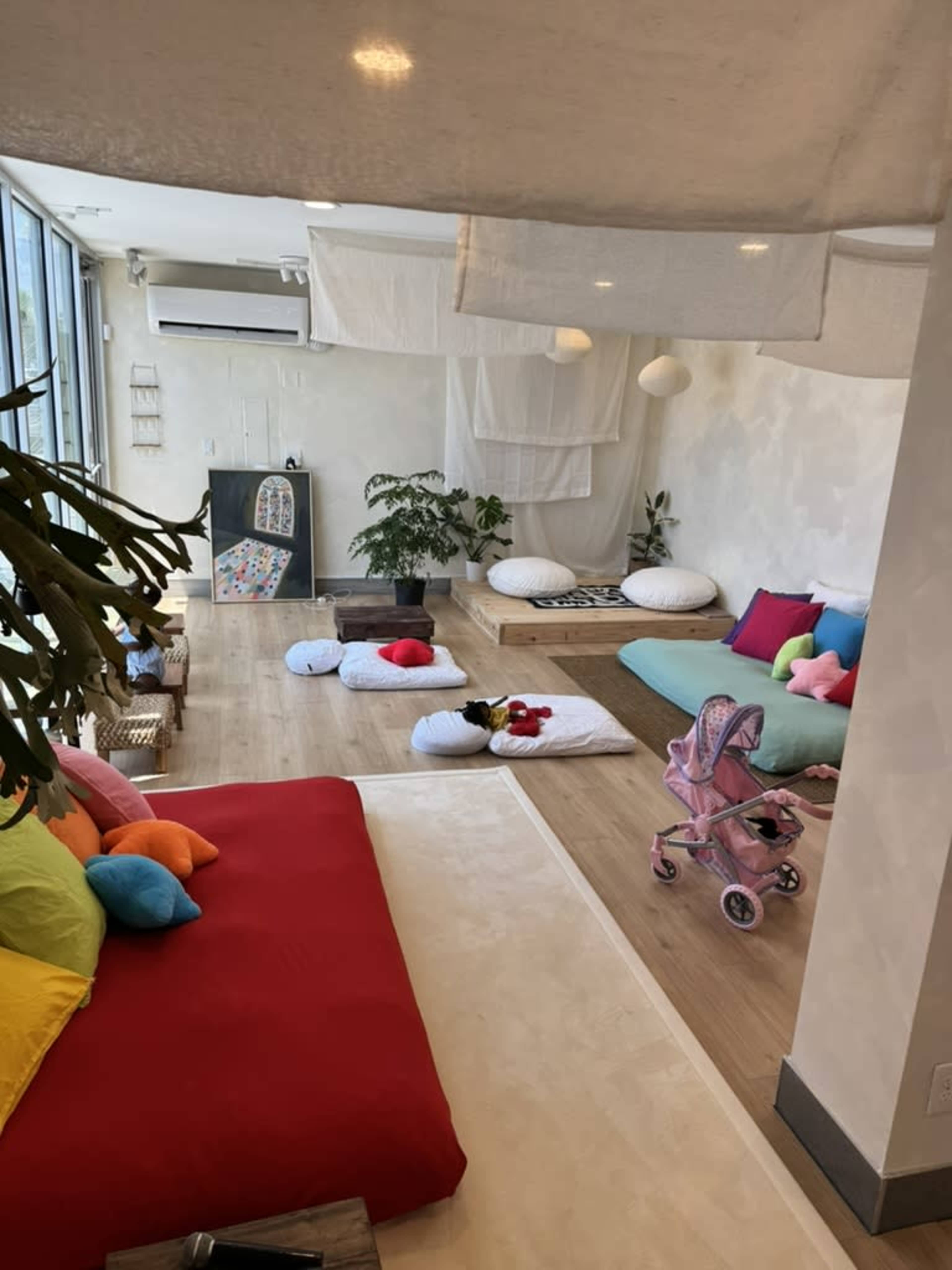 The image shows a cozy, open space with colorful cushions, a stroller, and plants, designed for relaxation and play.