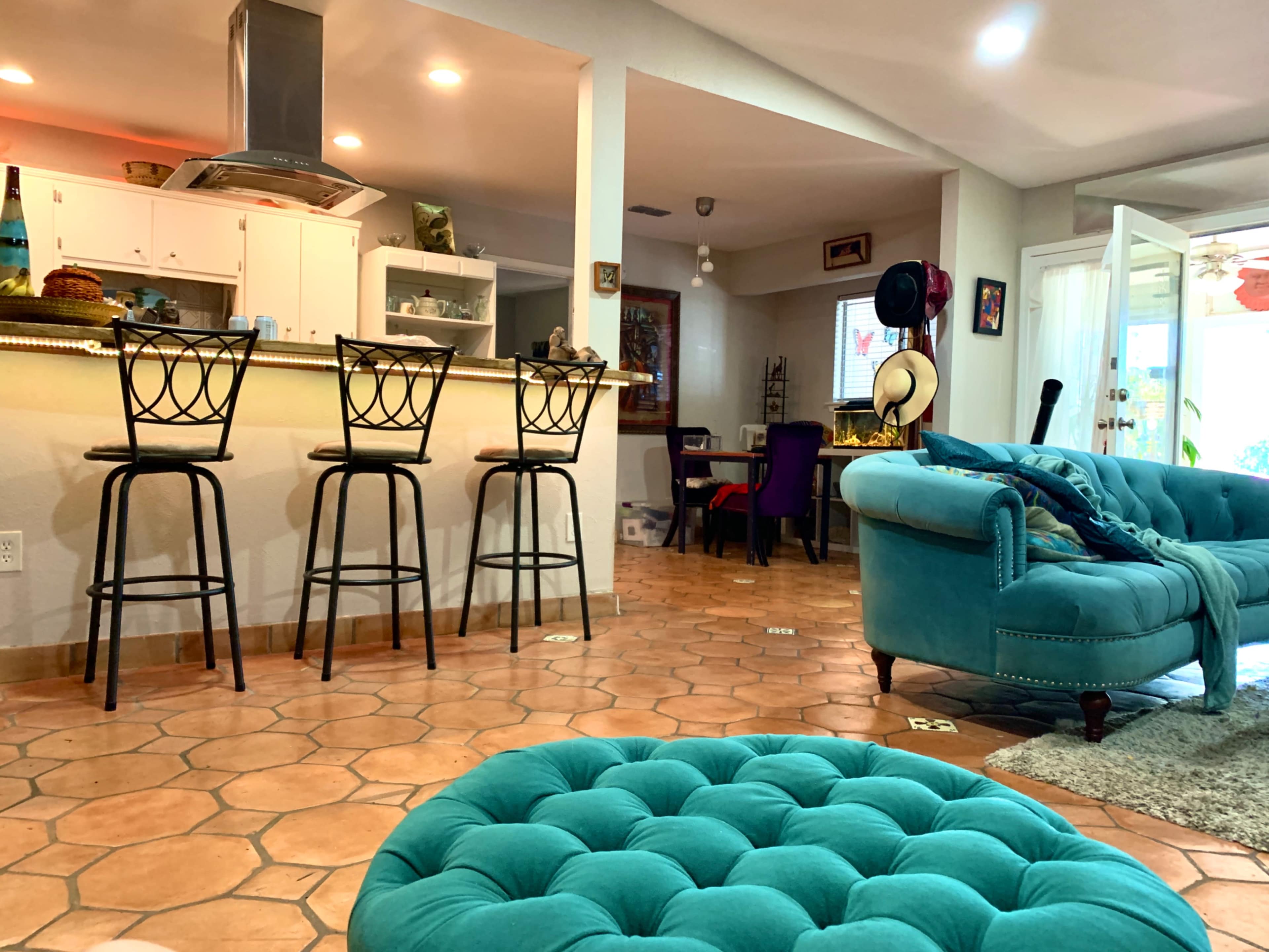 A living area featuring a turquoise sofa, a round ottoman, and a kitchen bar with four high stools.