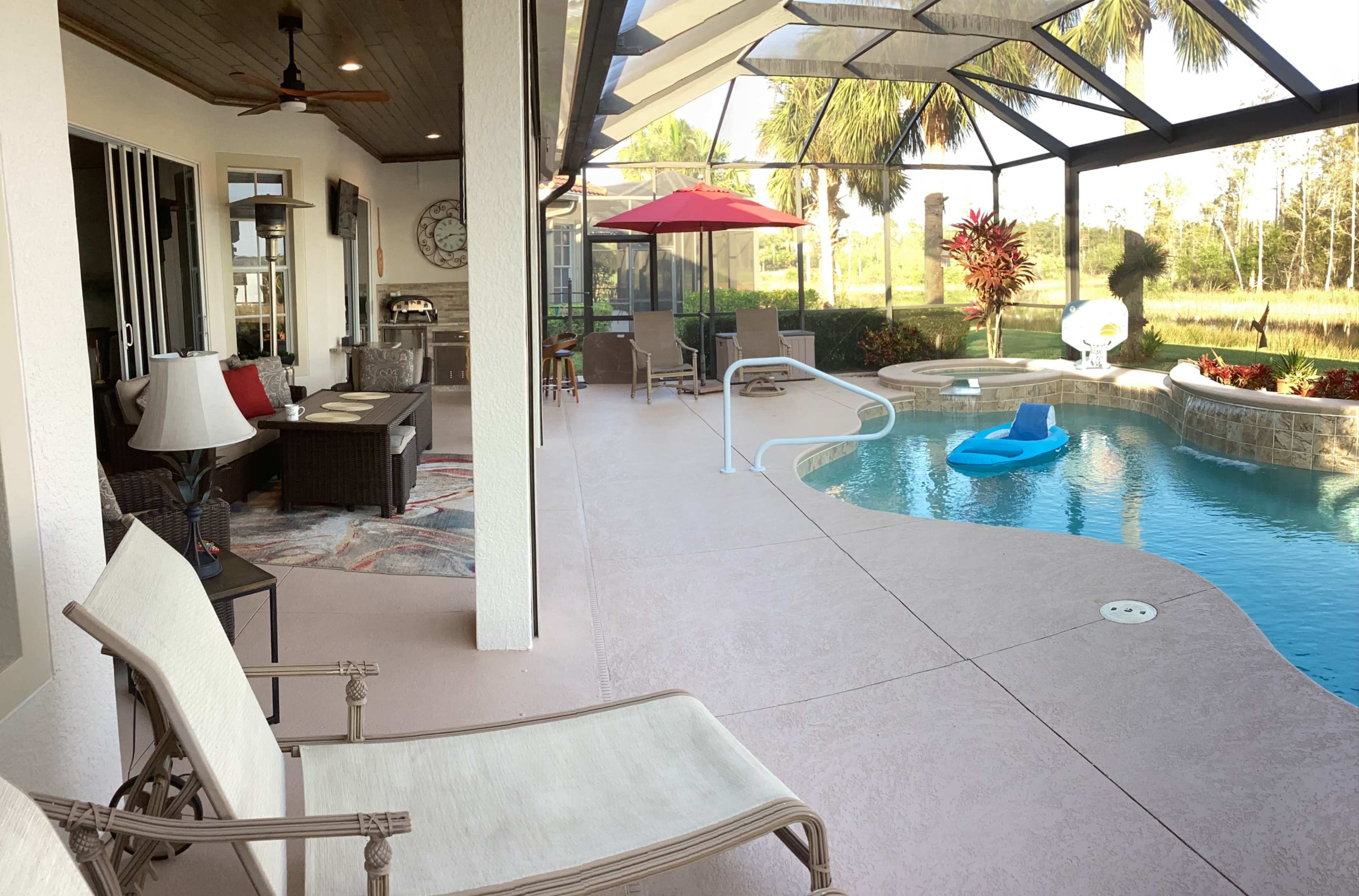A patio area beside a swimming pool with lounge chairs, a small table, and a red umbrella in a landscaped yard.
