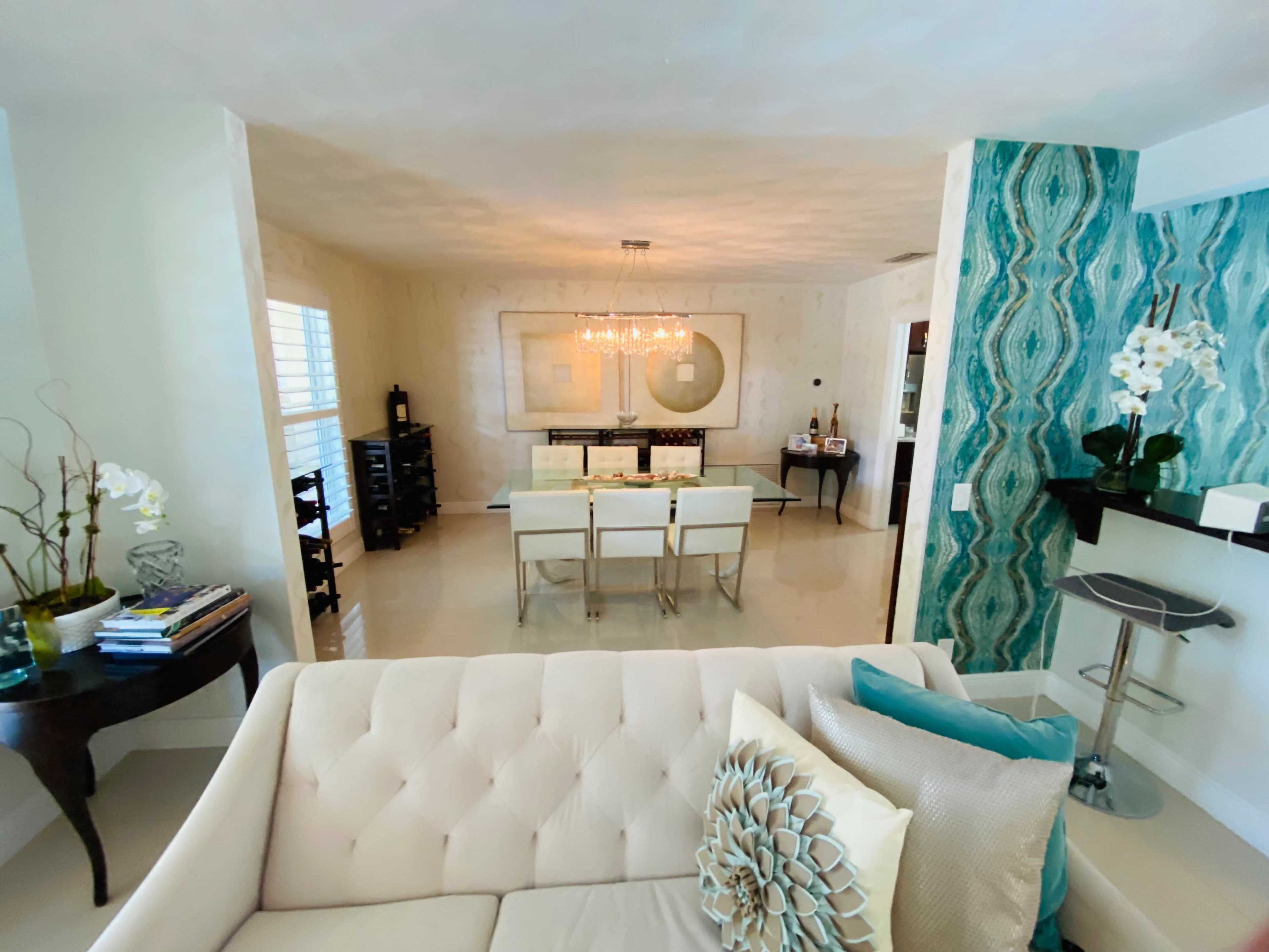 A modern living area features a light-colored sofa with decorative cushions, a dining space with a table and chairs, and walls decorated with a textured teal design.