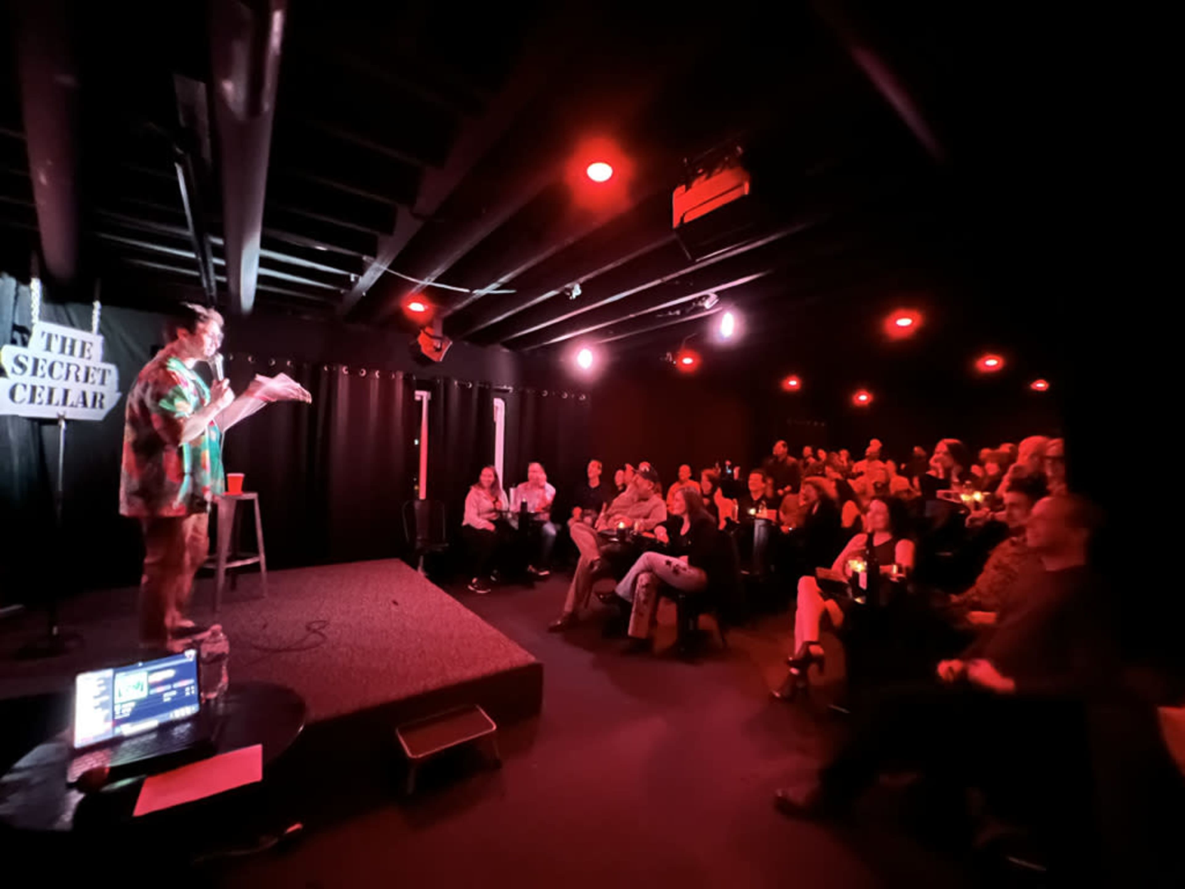 A performer stands on stage in a dimly lit venue with red lighting, addressing an audience of seated viewers.
