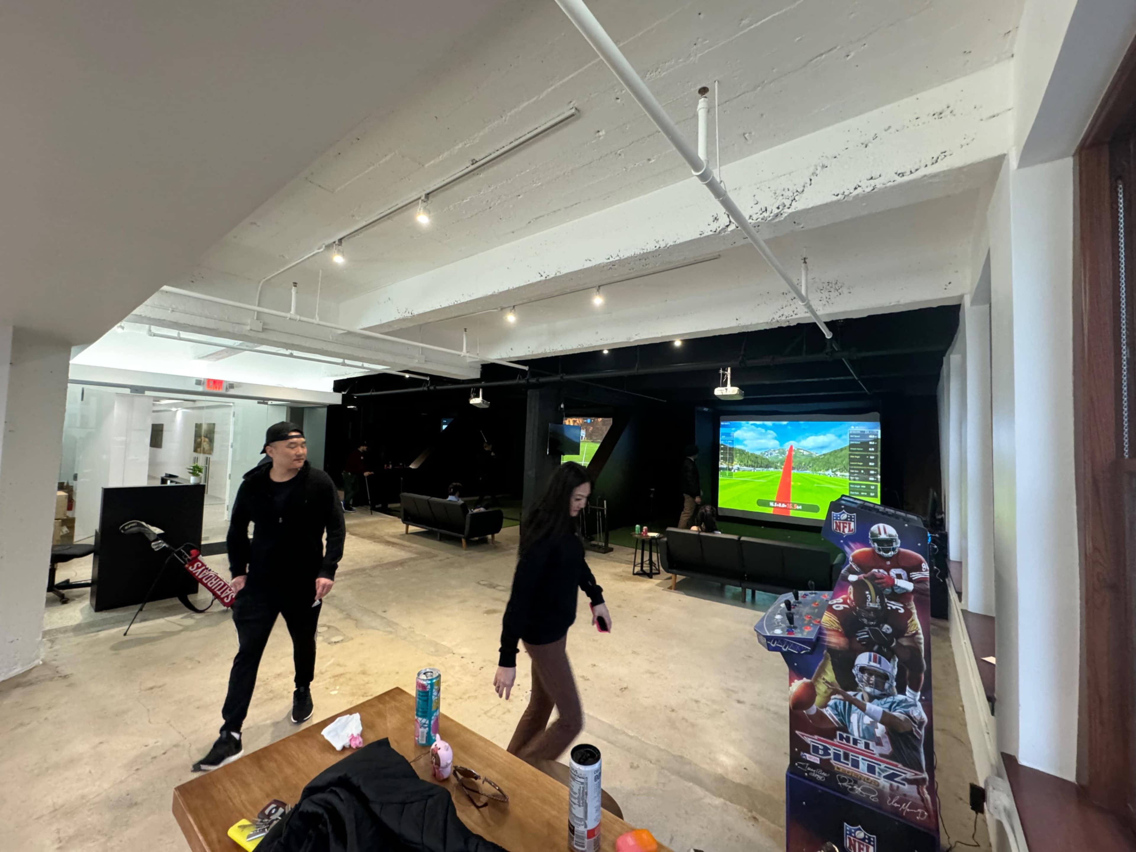 A casual lounge area features two people walking past a large TV displaying a sports game, with seating and gaming equipment visible in the background.