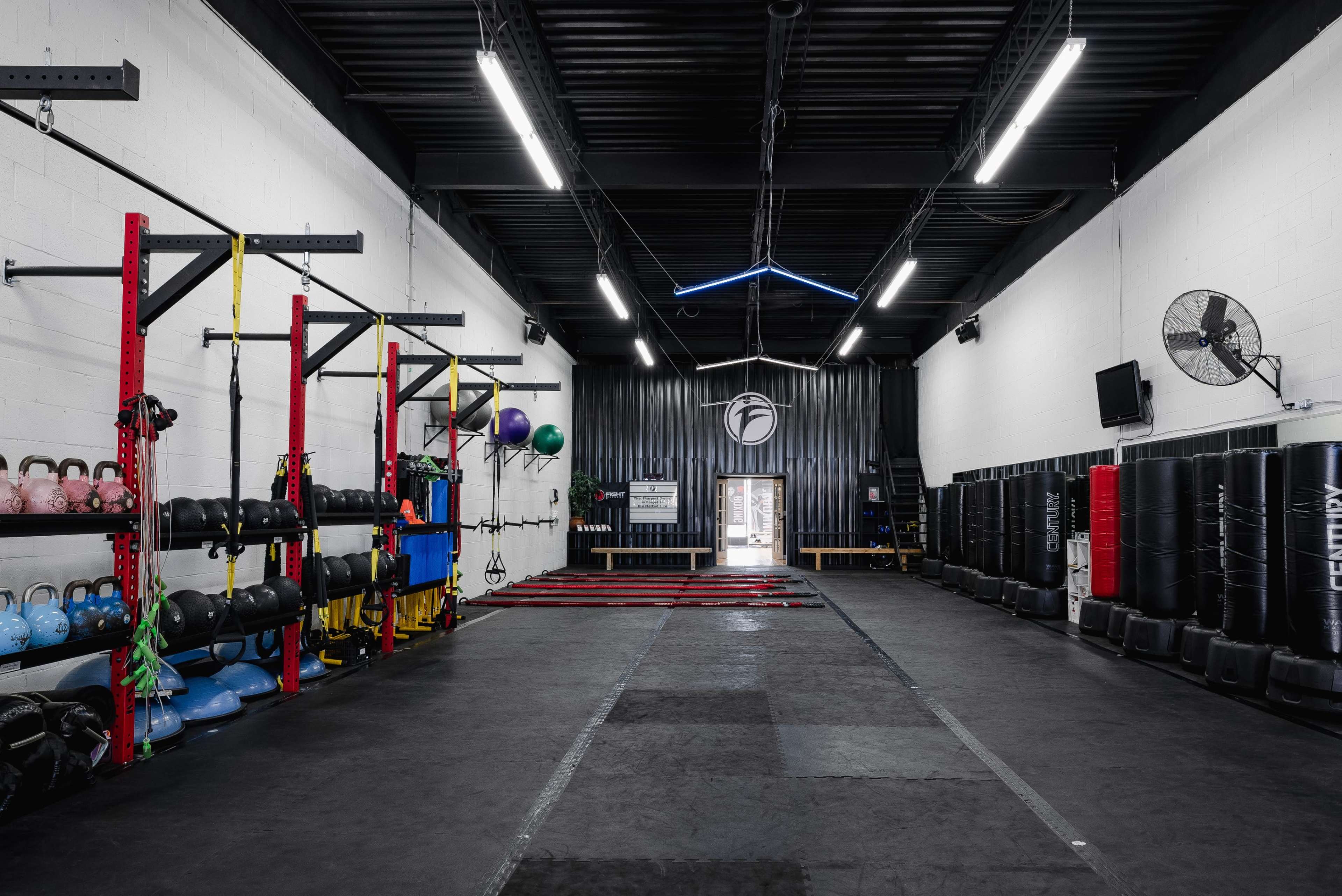 A spacious gym interior with exercise equipment, including kettlebells, resistance bands, and punching bags, organized along the walls.