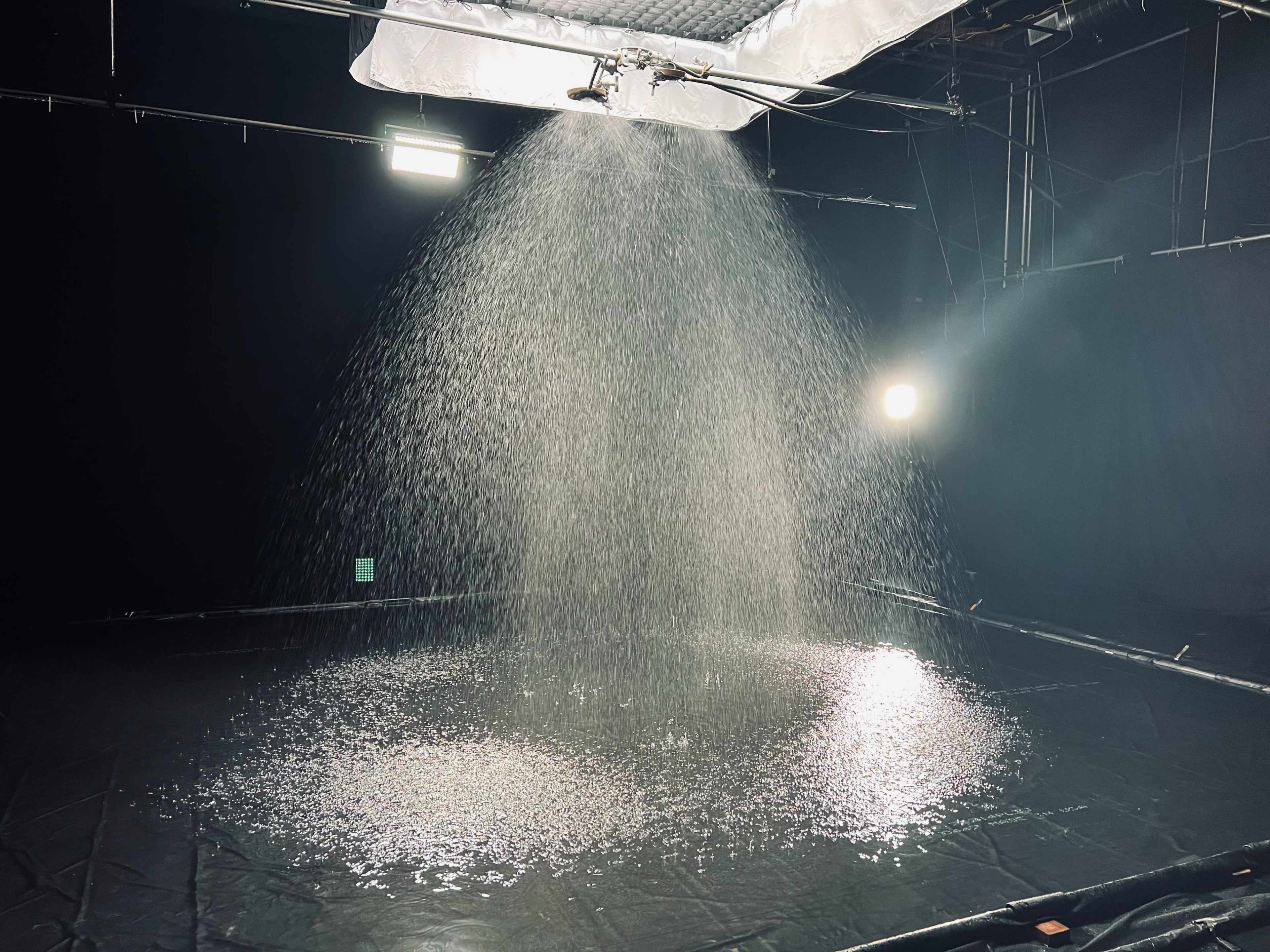 A water spray system creates a circular pool of water on a black surface in a dimly lit studio.