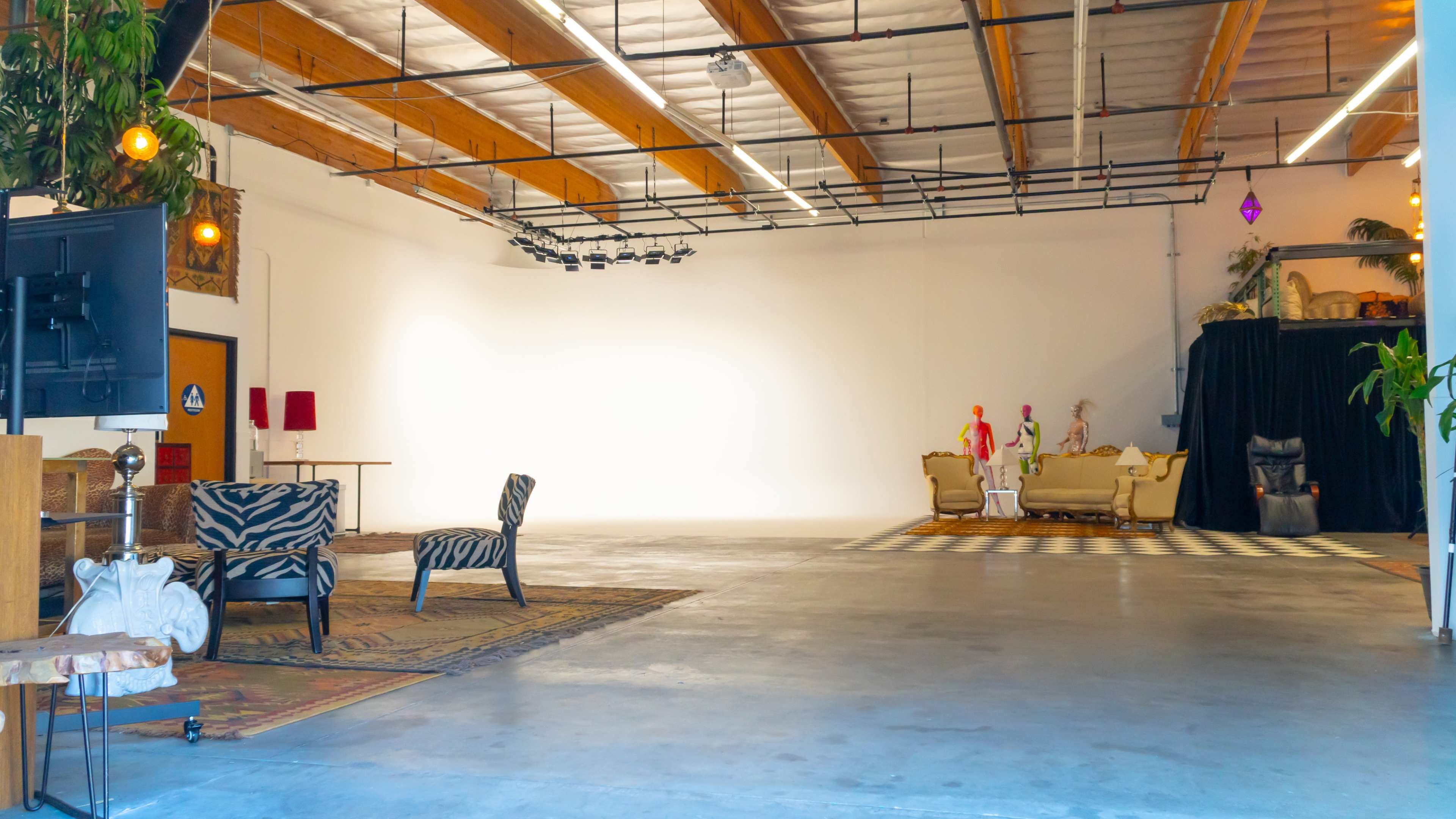 A spacious, well-lit studio with wooden beams, two mannequins positioned near a seating area, and a variety of furniture and decor items scattered throughout the concrete floor.