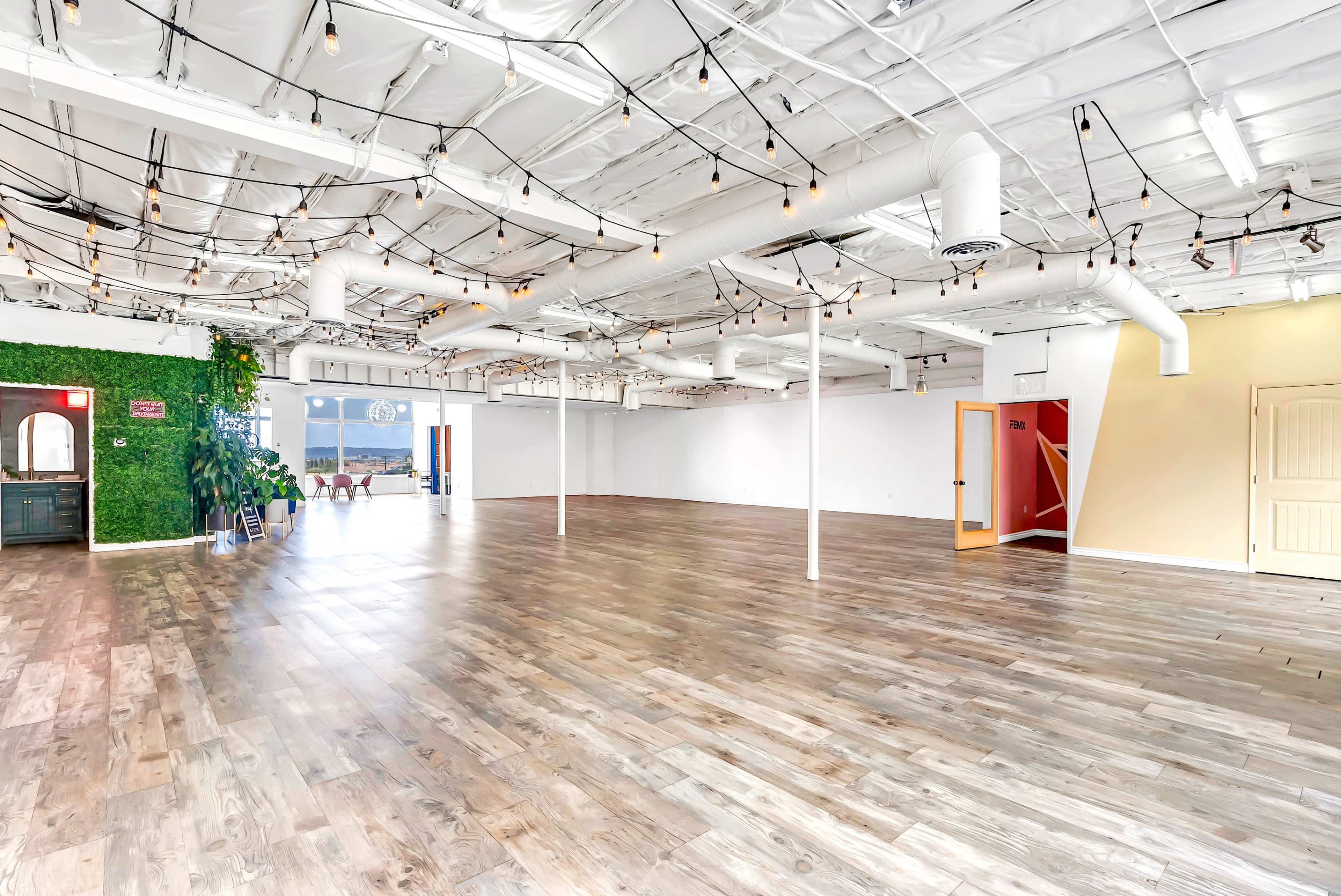 A spacious, empty room with wooden floors, white walls, and exposed ceiling beams adorned with hanging string lights.