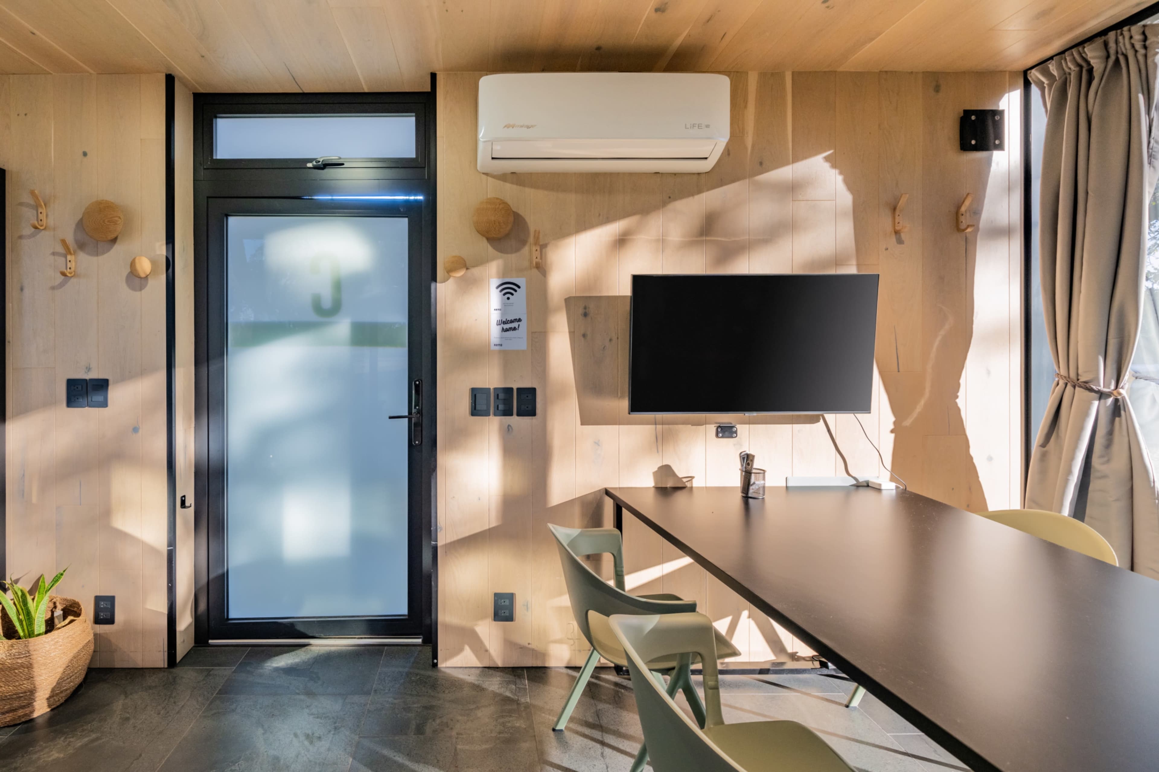 The image shows a modern room featuring a wooden interior with a large table, several chairs, a television mounted on the wall, and a door marked with the number 3.