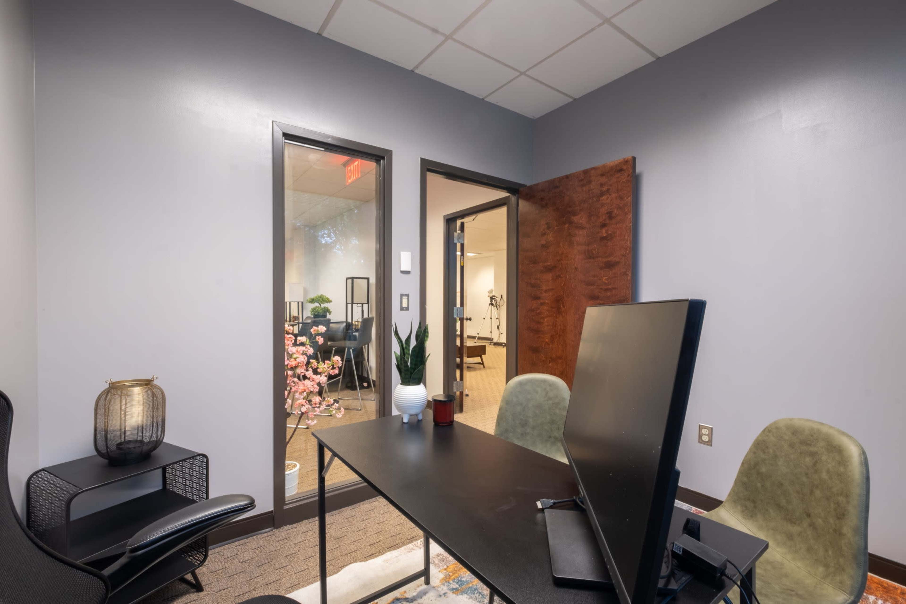 The image shows a small office interior with a black desk, a monitor, two chairs, and large windows allowing light to enter from an adjacent room.