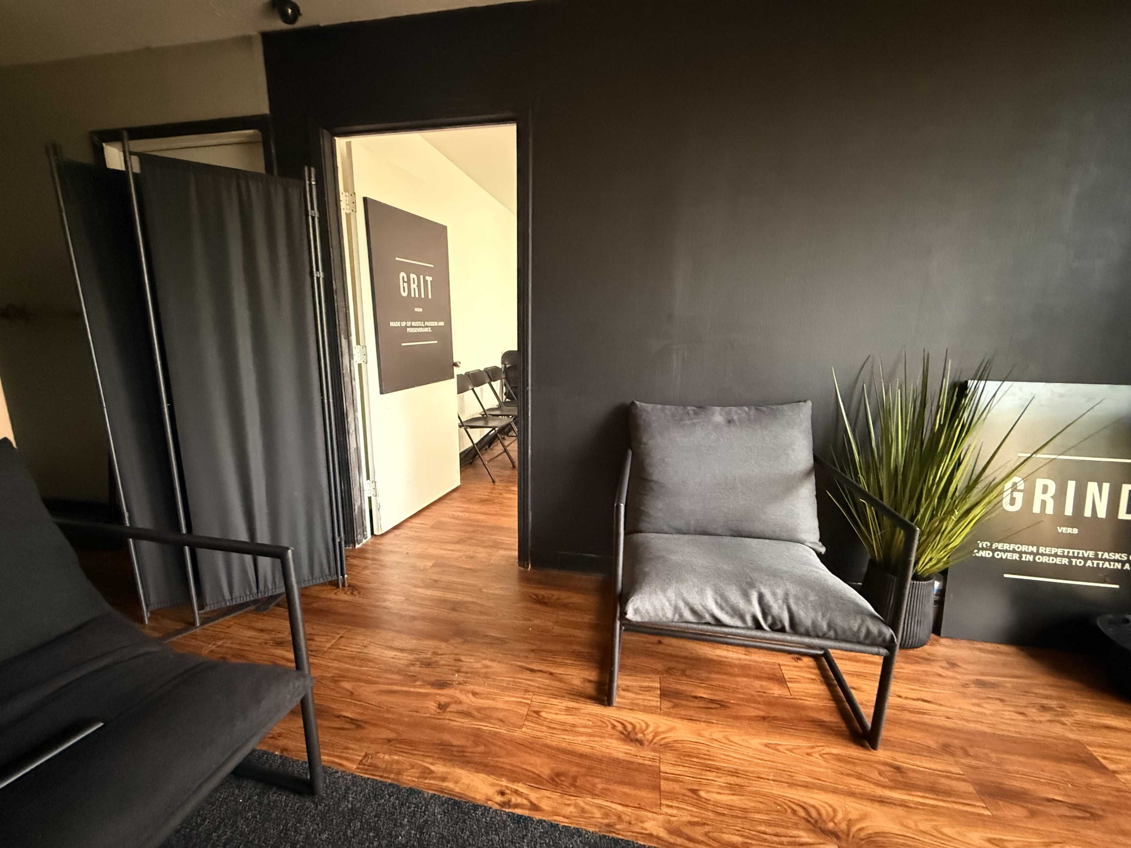 The image shows a modern waiting area with two gray chairs, a plant, and a doorway leading to another room with a sign that reads "GRIT."