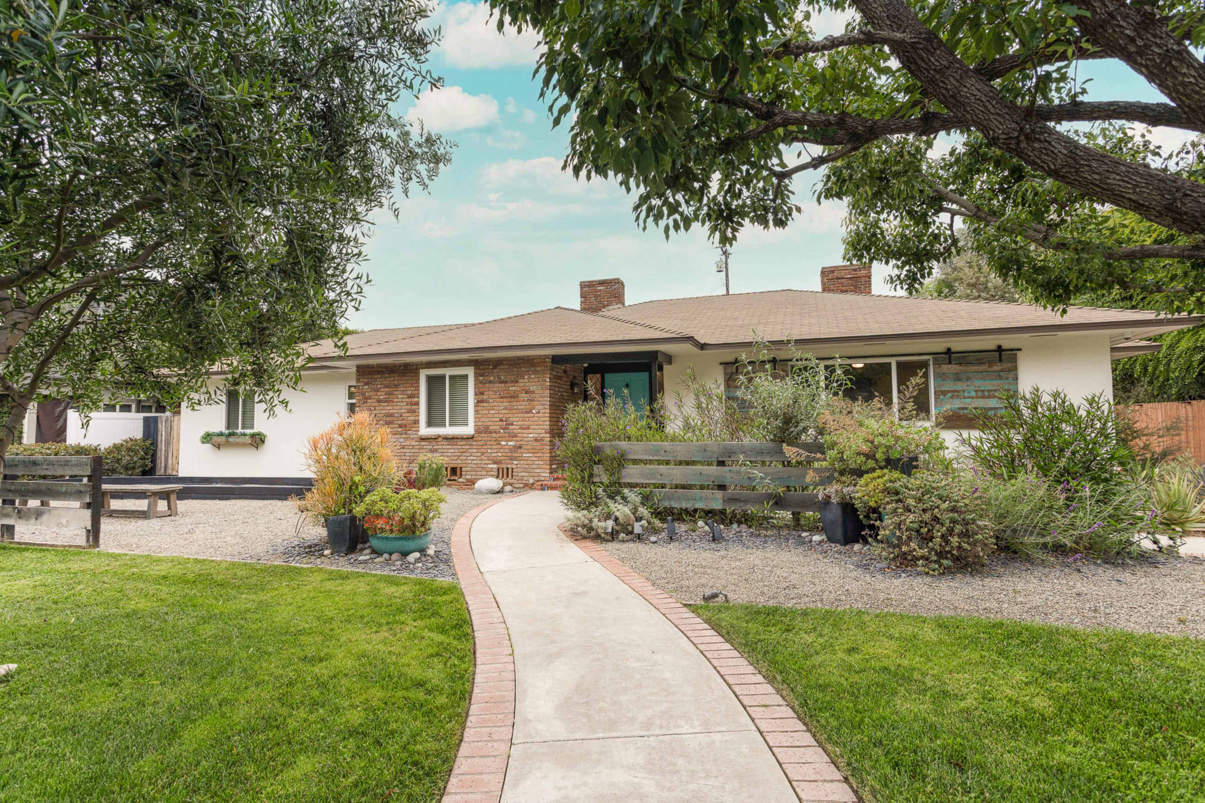 A single-story house with a brick accent wall is surrounded by various plants and a curved pathway leading to the front door.