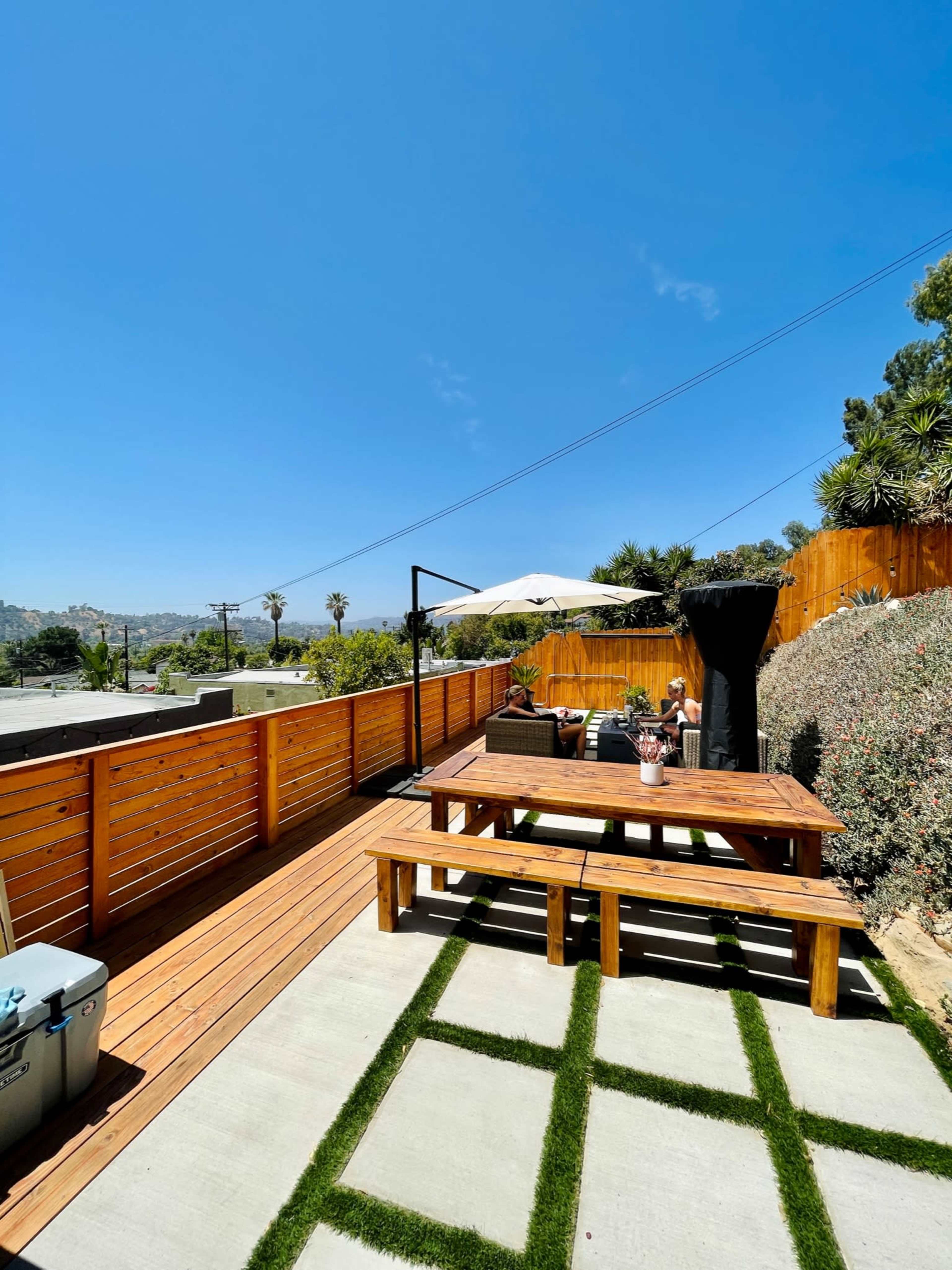 The image shows a wooden deck with a large table and an umbrella, surrounded by greenery and a concrete pathway under a clear blue sky.