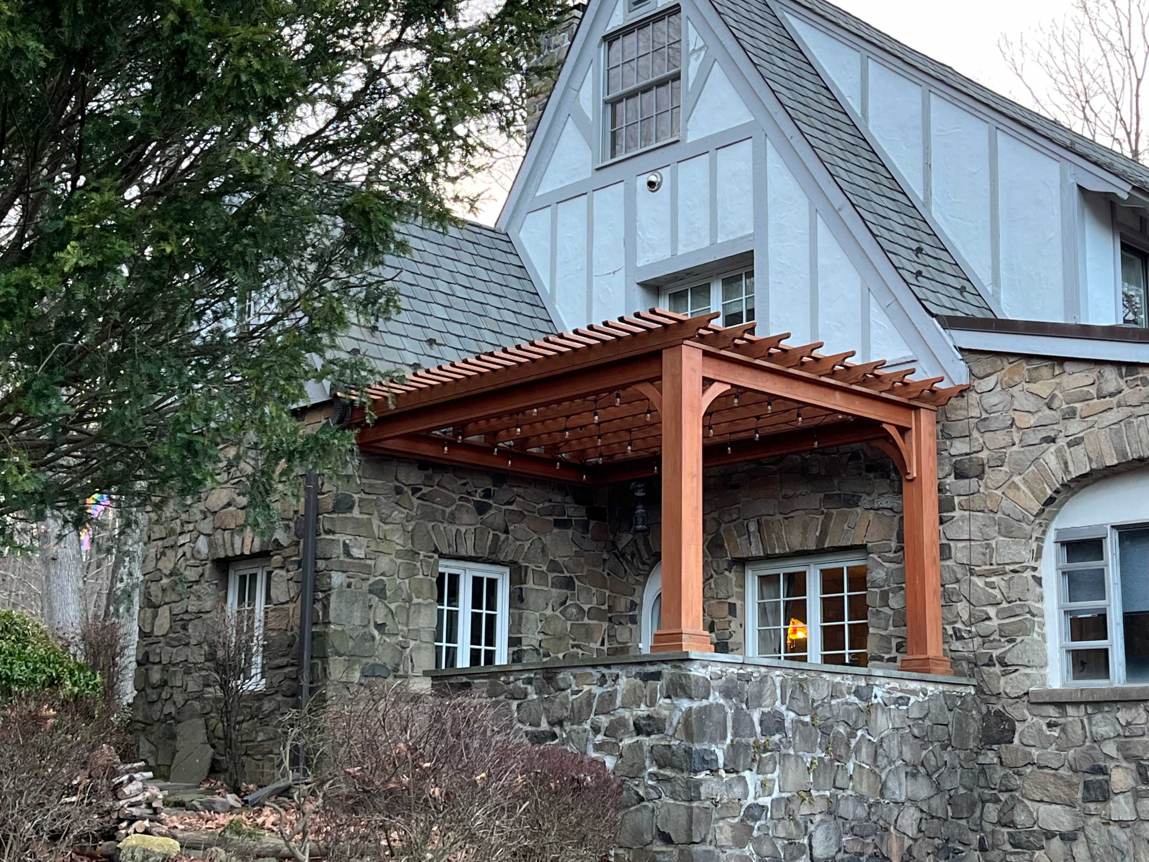 The image shows a stone-clad house with a wooden pergola overhanging a patio area.