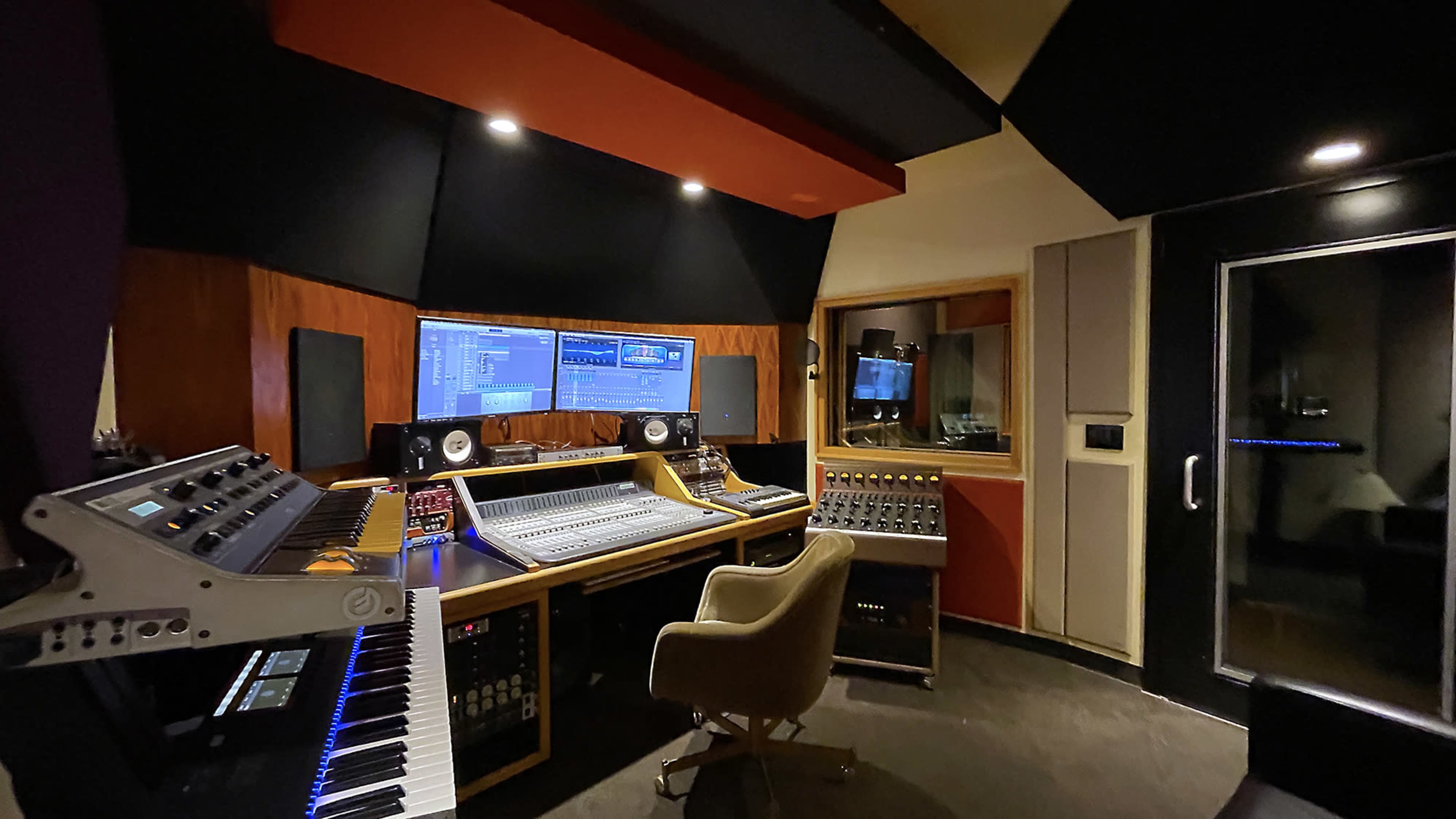 The image shows a modern recording studio featuring a large sound mixing console, multiple computer screens, studio monitors, and a keyboard.