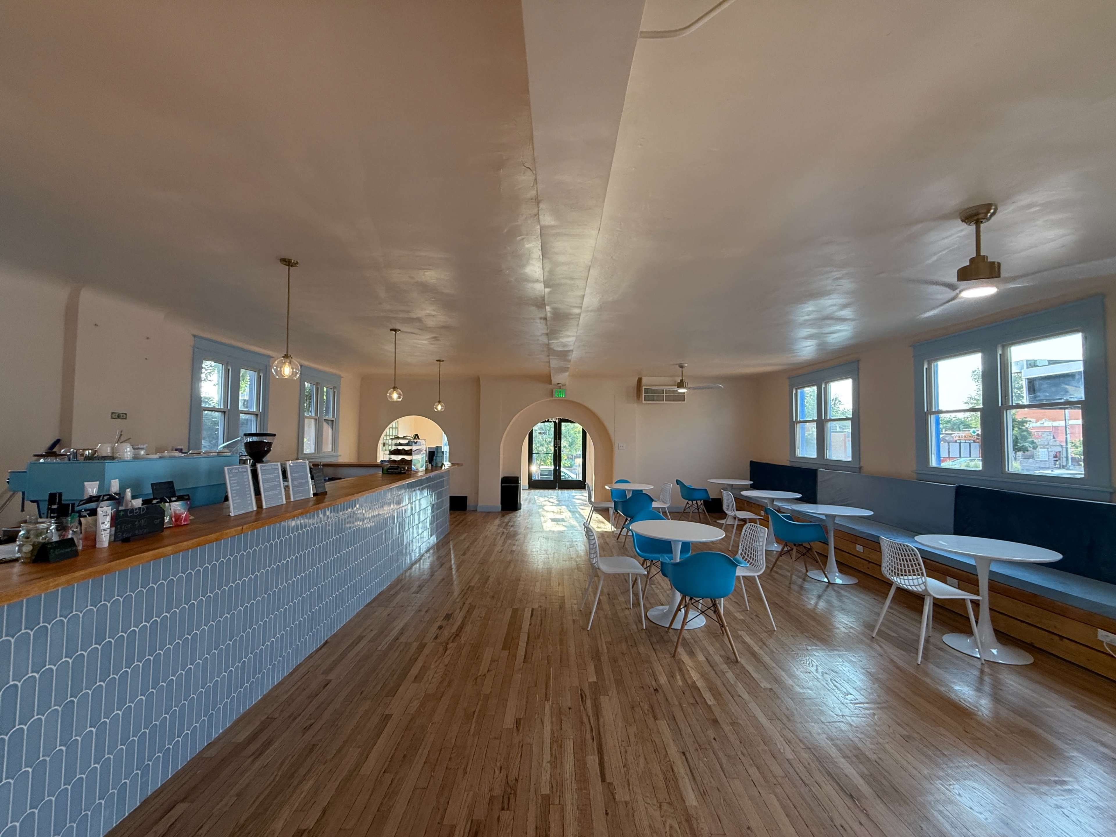 The image shows a modern, light-filled café interior with a wooden floor, blue tile countertop, and various tables and chairs arranged for customers.