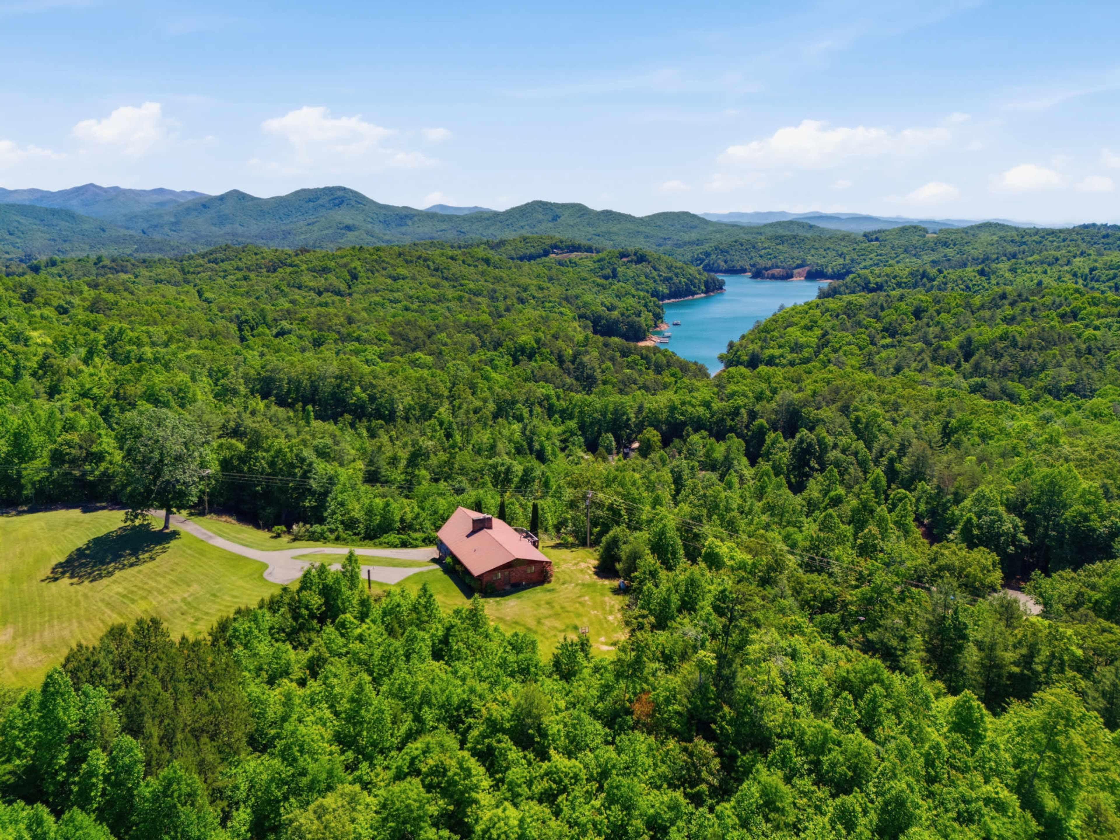 A large house with a red roof sits surrounded by dense greenery, next to a tranquil blue lake and distant mountains.