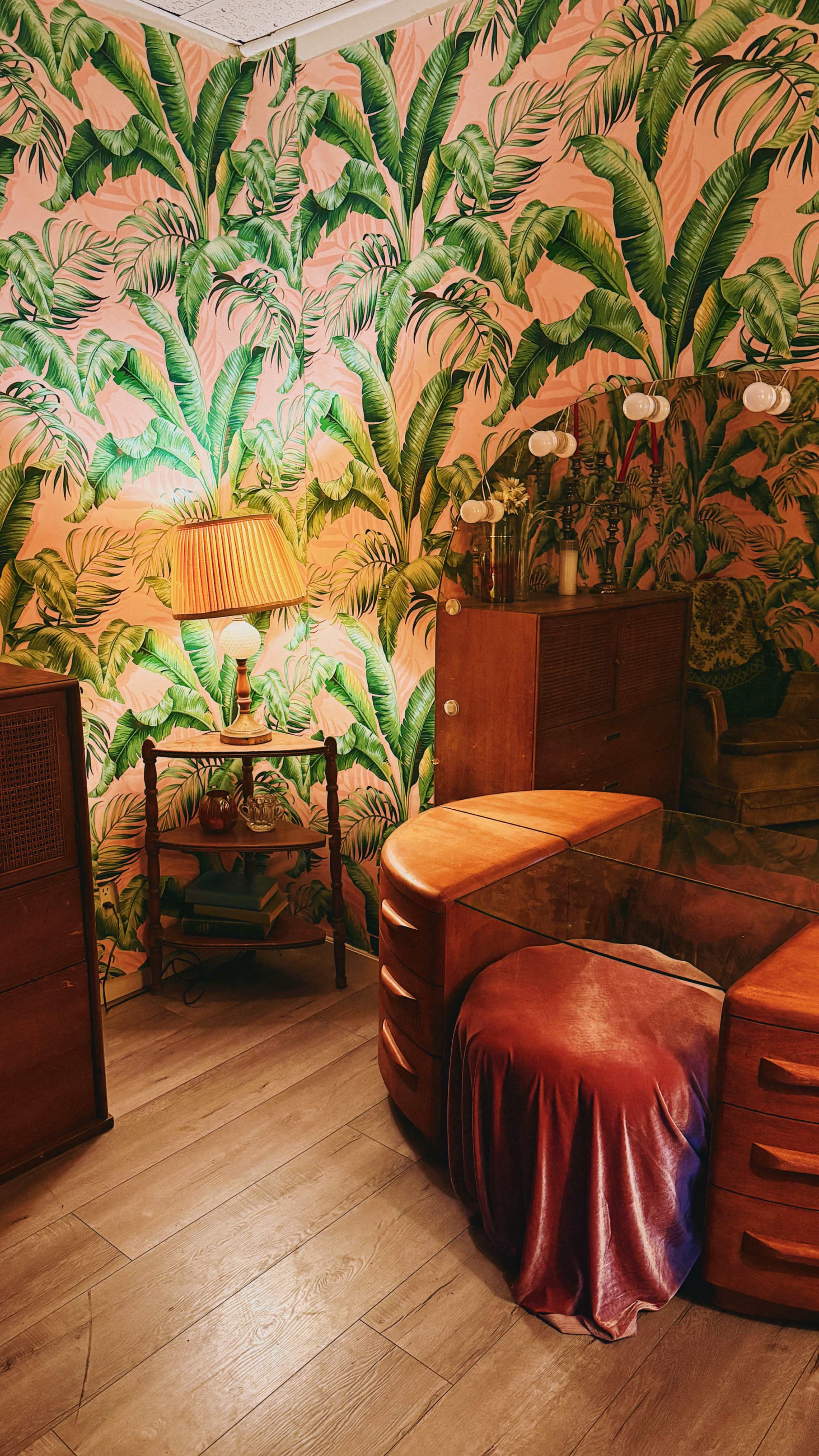 The image depicts a room with tropical-print wallpaper, featuring a curved vanity with a round stool, a small wooden side table, and a lamp.