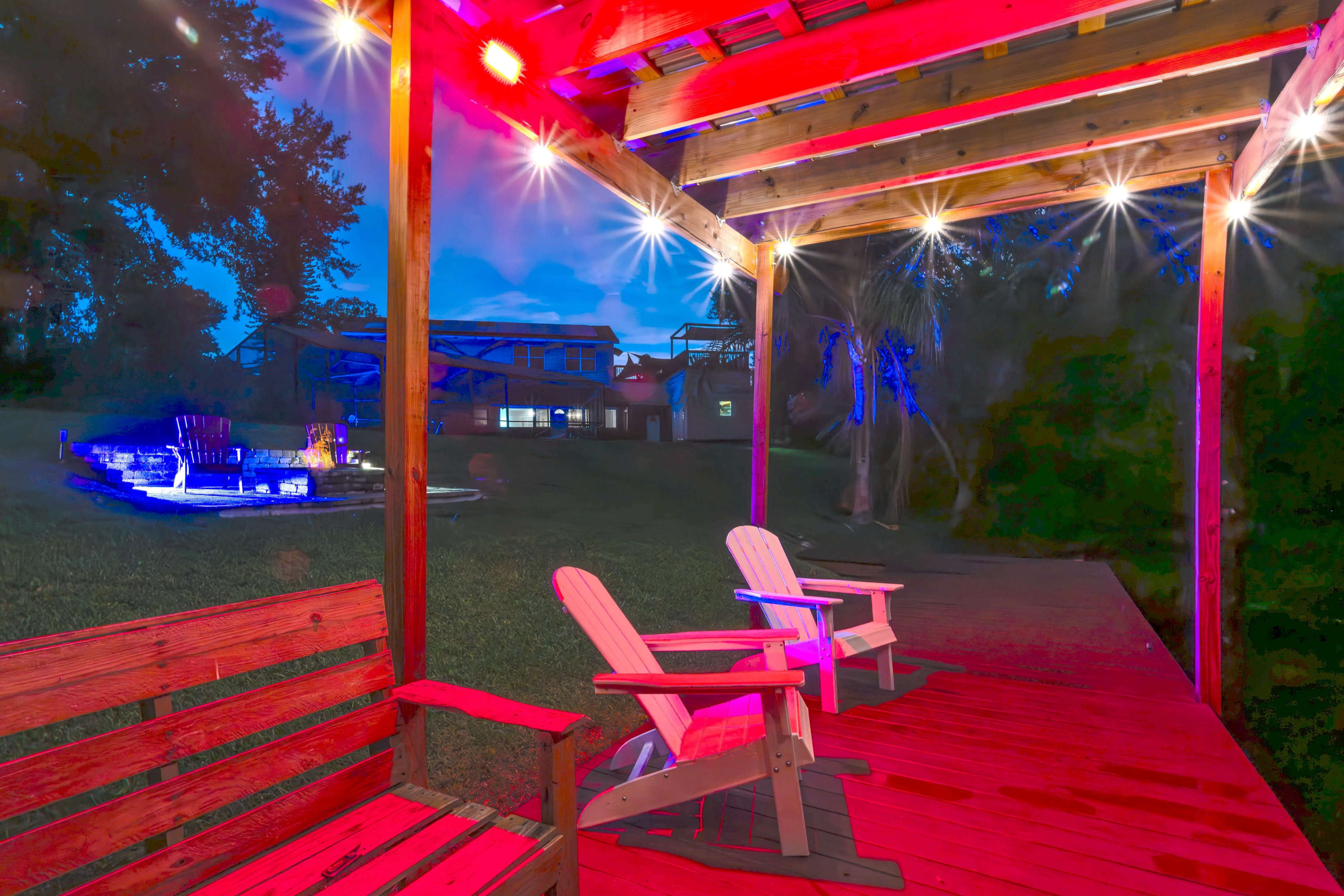 A wooden deck with two Adirondack chairs is illuminated by colored lights beneath a covered structure, while the background features a grassy area and a lit pool.
