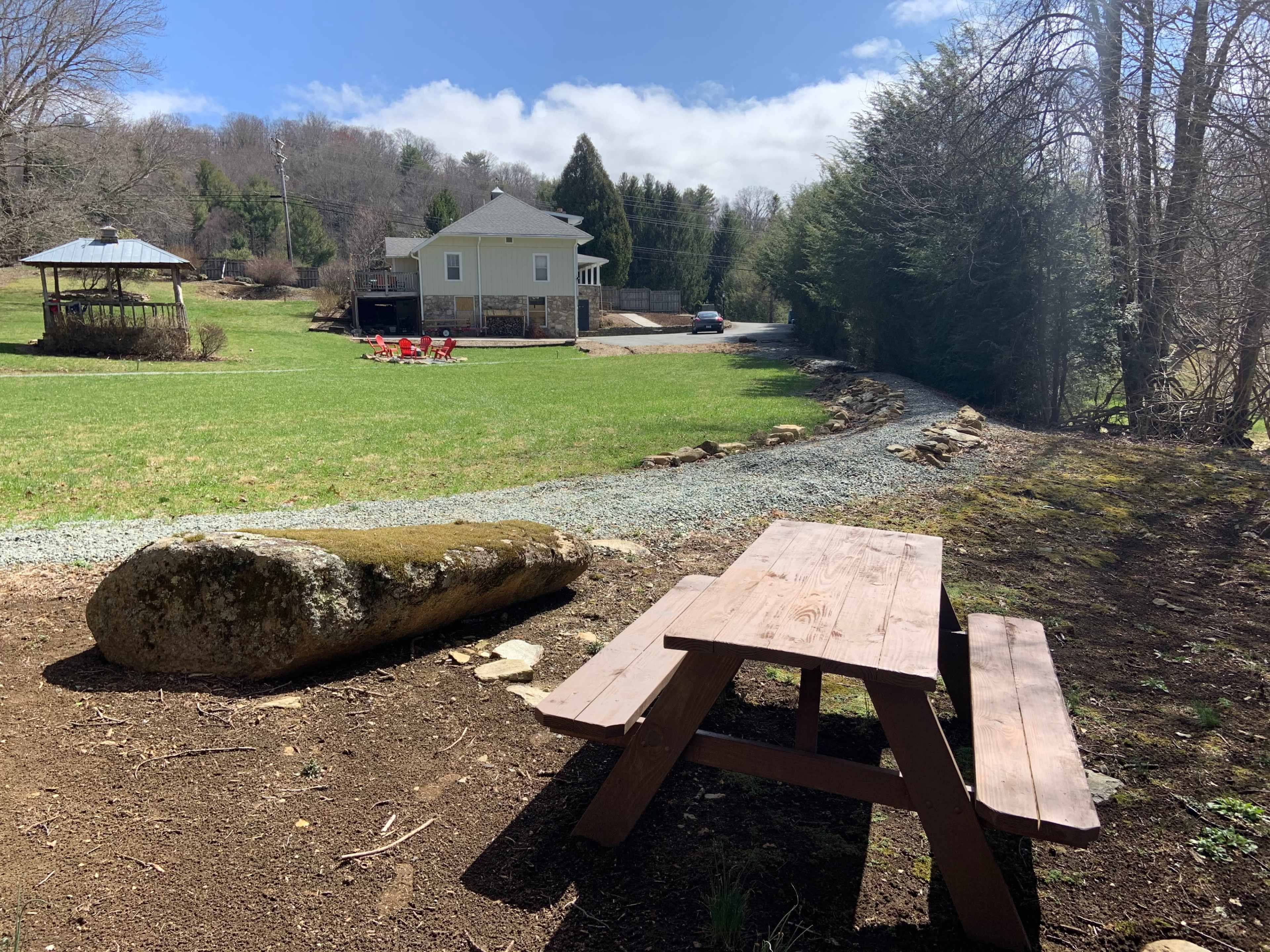 A wooden picnic table sits on a dirt area near a grassy lawn, with a stone and gravel path leading to a house and gazebo in the background.