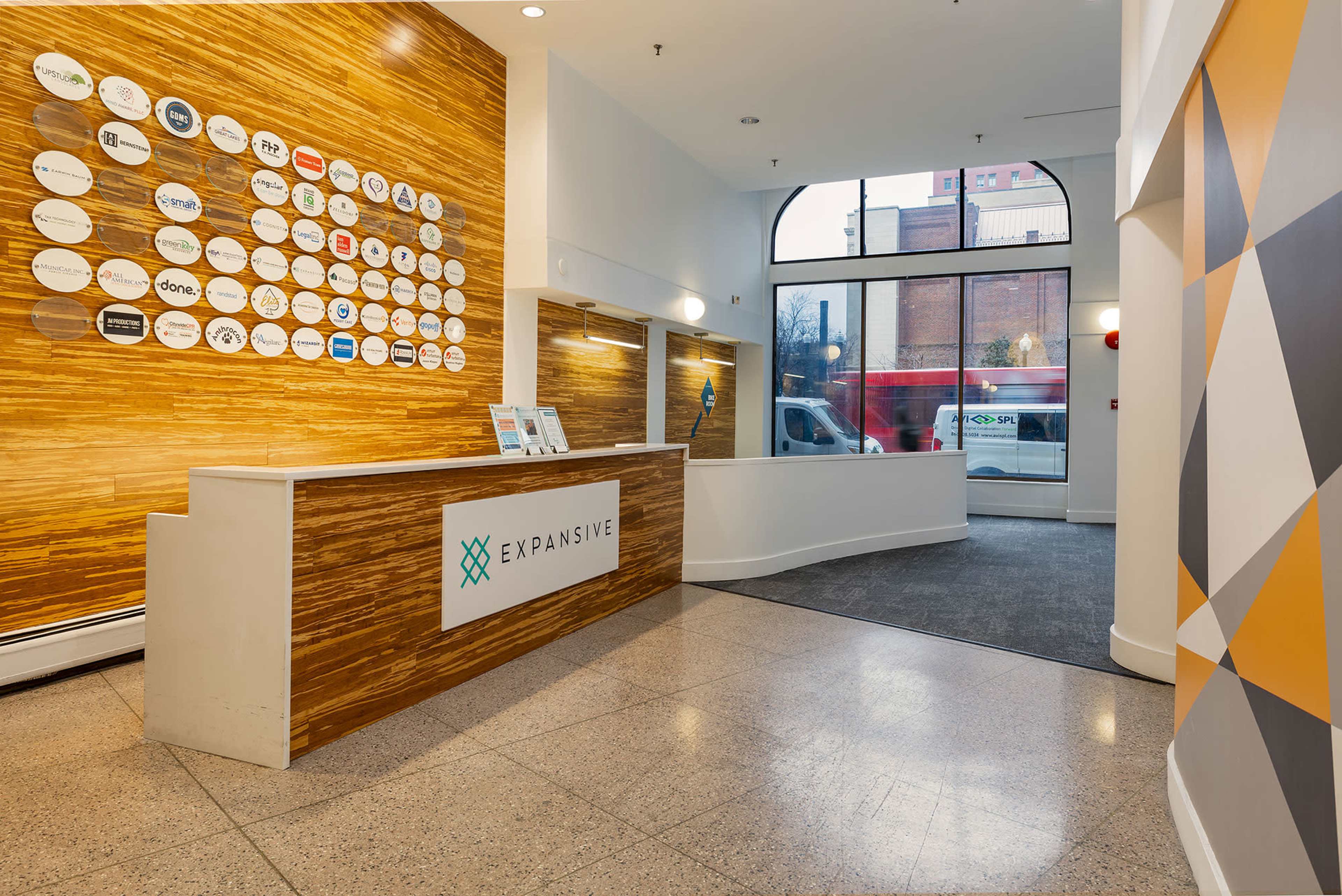 The image shows a modern reception area featuring a wooden accent wall with various logos, a sleek front desk with the "EXPANSIVE" sign, and a large window allowing natural light into the space.