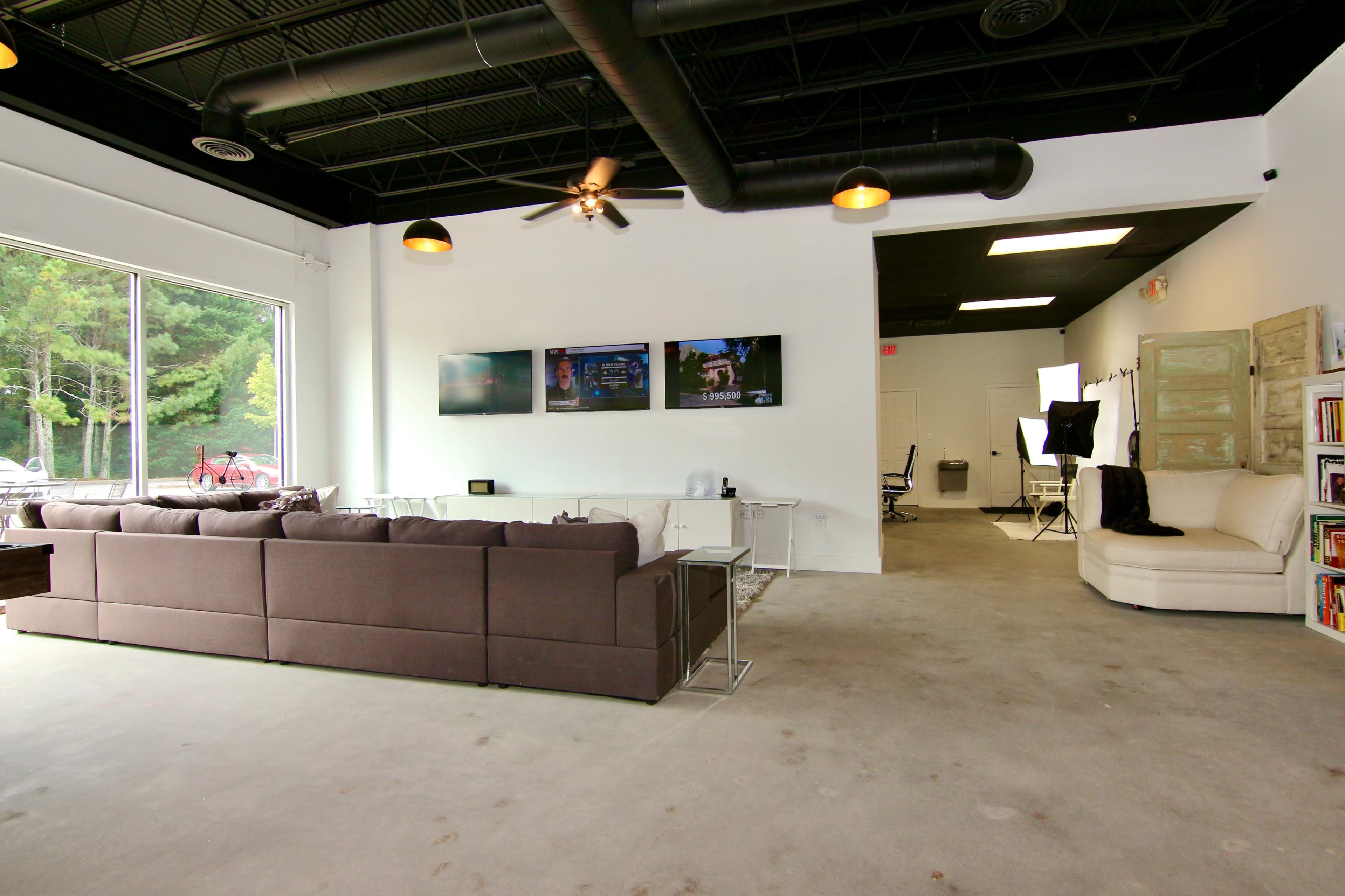 A modern open space with a gray sectional sofa, three wall-mounted screens, and a seating area in a well-lit room featuring large windows.