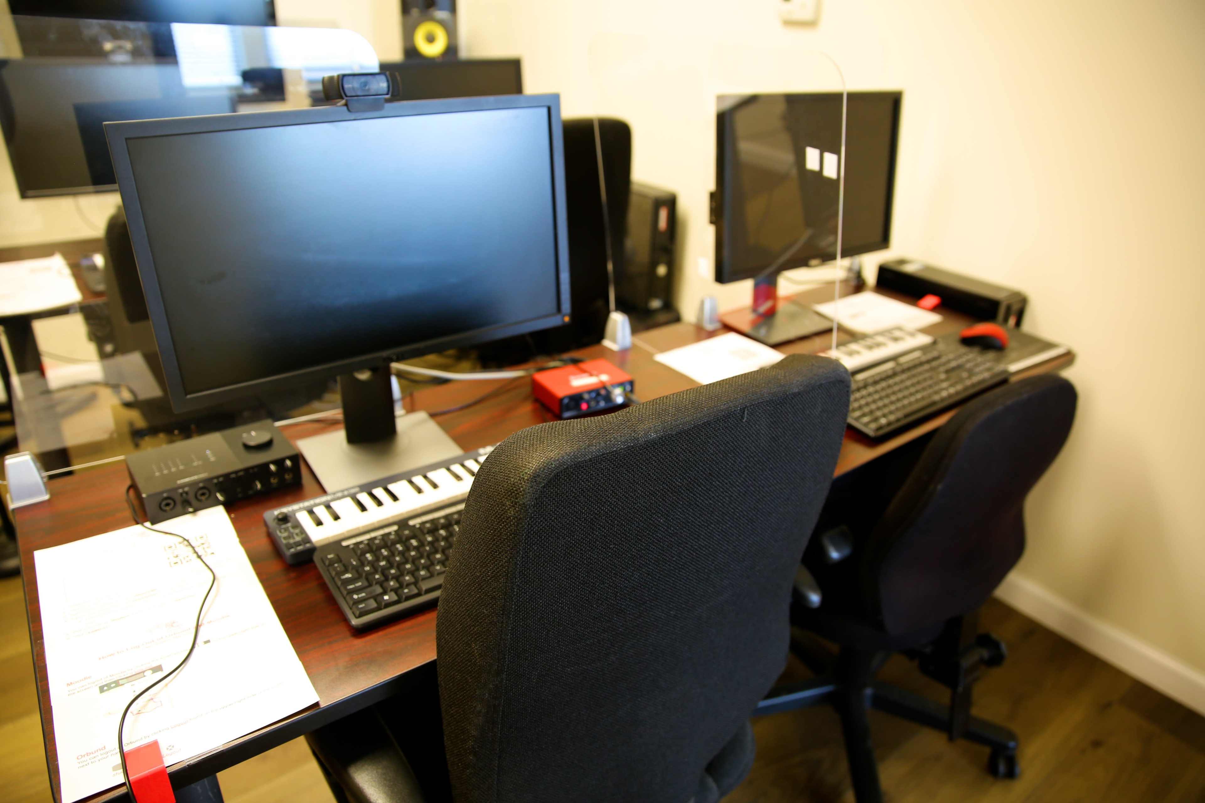The image shows two computer workstations with monitors, keyboards, and audio equipment in an office setting.