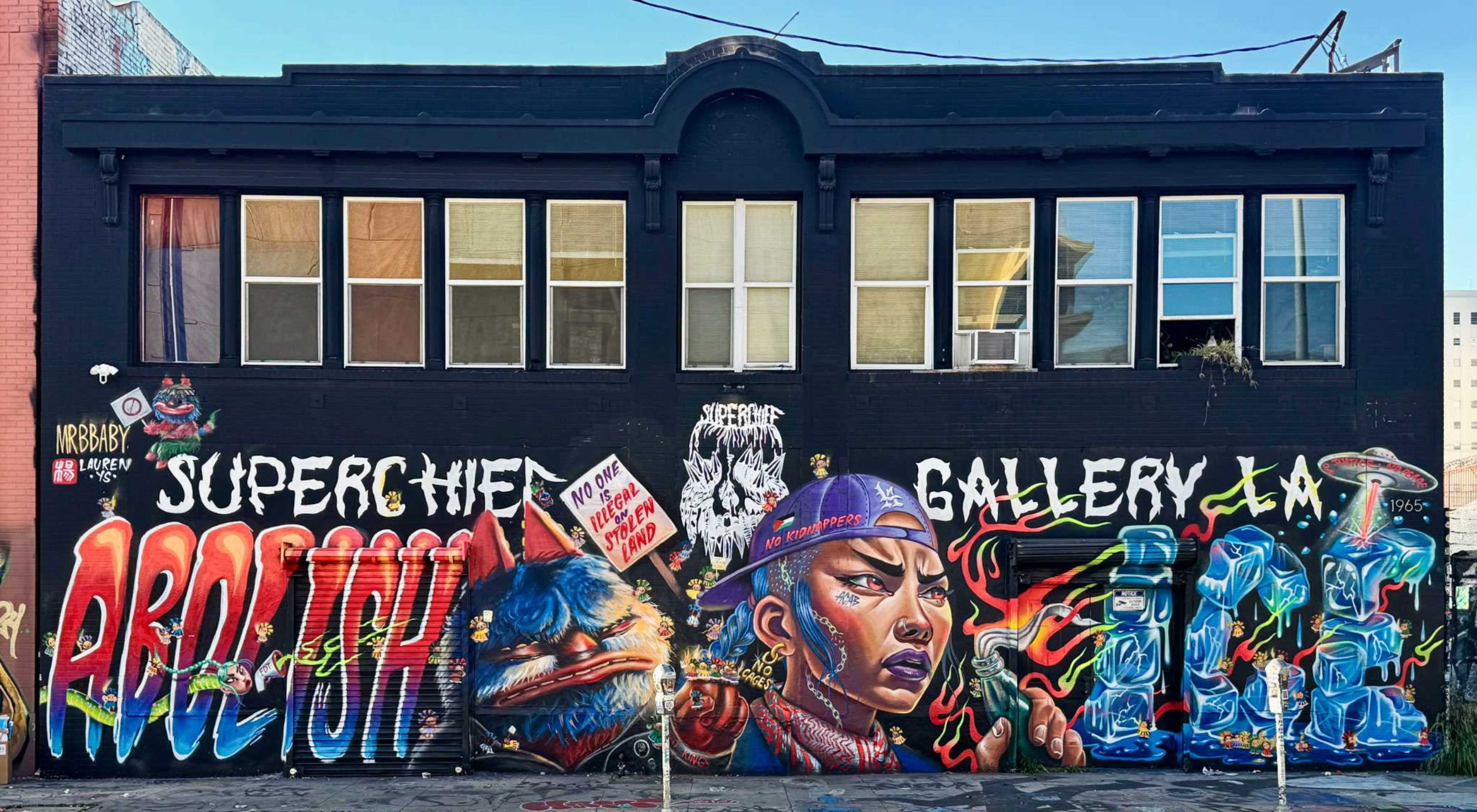 The image shows a large mural on a black building wall featuring vibrant graffiti art, including the words "SUPERCHIEF" and "GALLERY," along with imagery of a woman's face and a stylized animal.