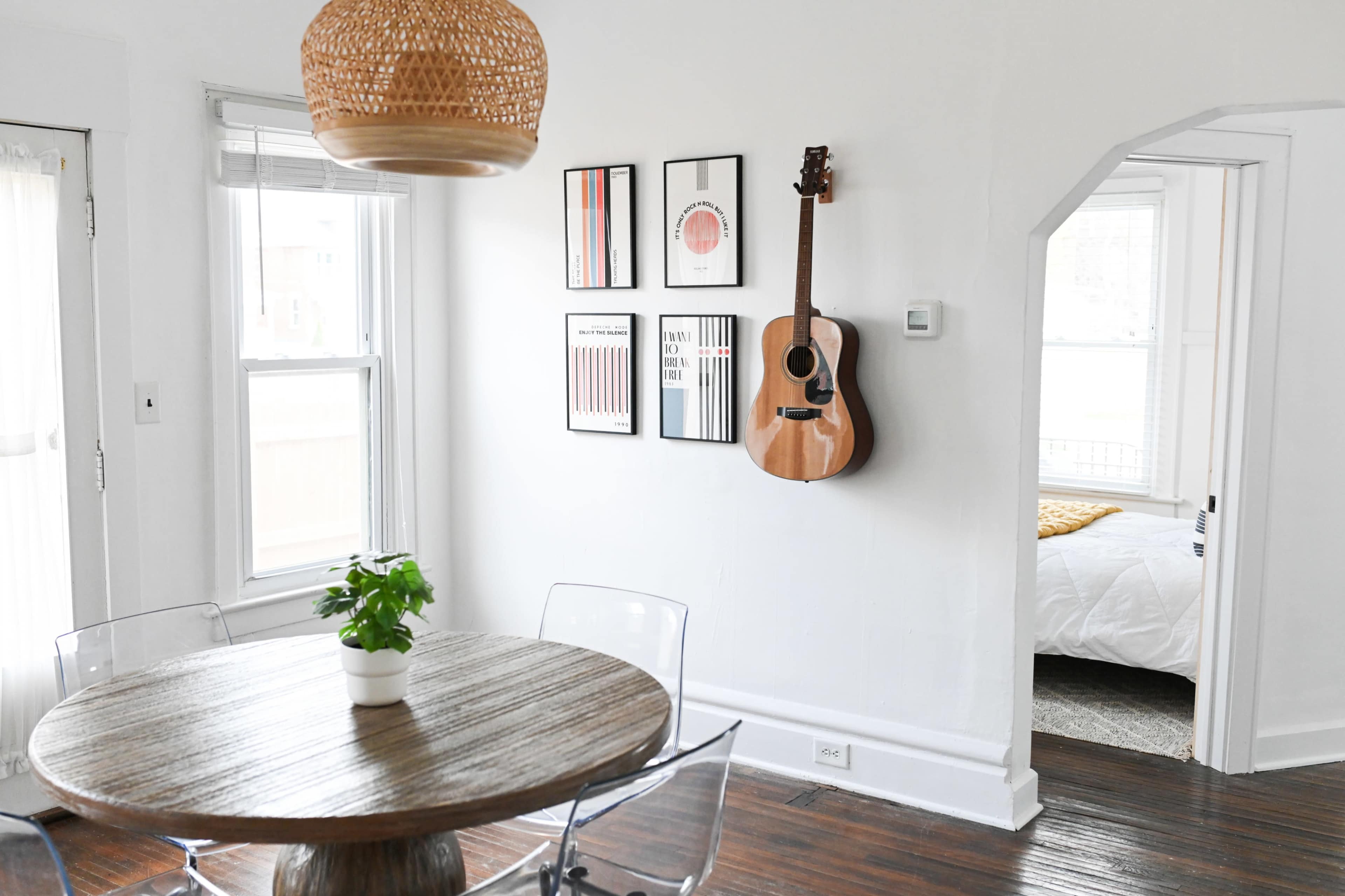 A round wooden dining table with clear chairs is positioned in a bright room featuring a decorative woven light fixture and a wall displaying framed prints and an acoustic guitar.