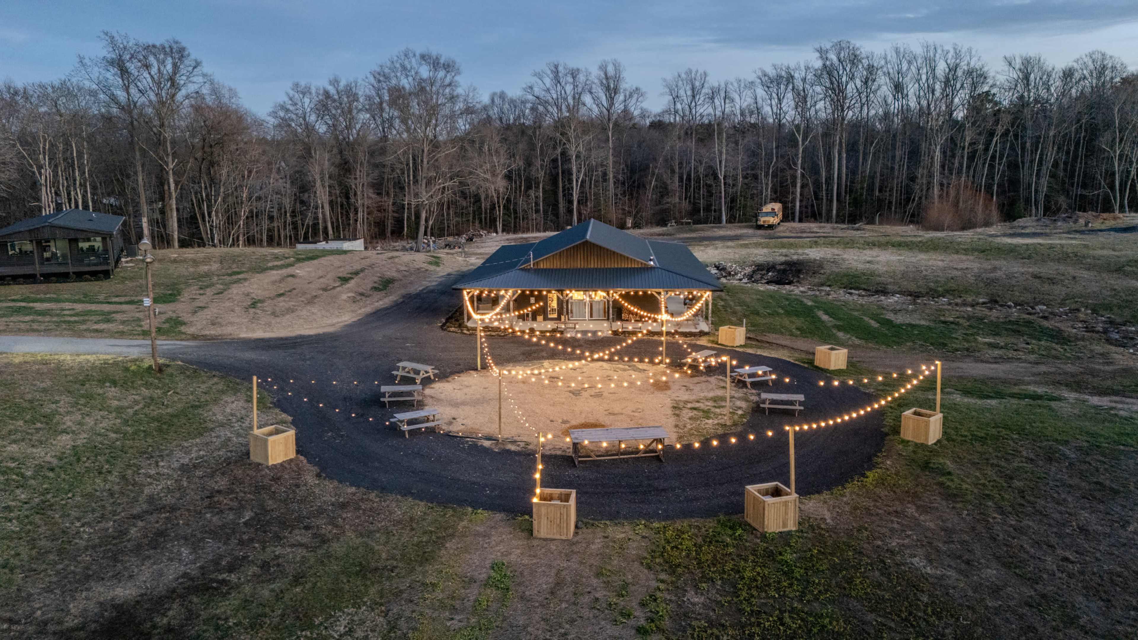 The image shows a circular gathering area with a wooden structure at the center, surrounded by picnic tables and lit by string lights.