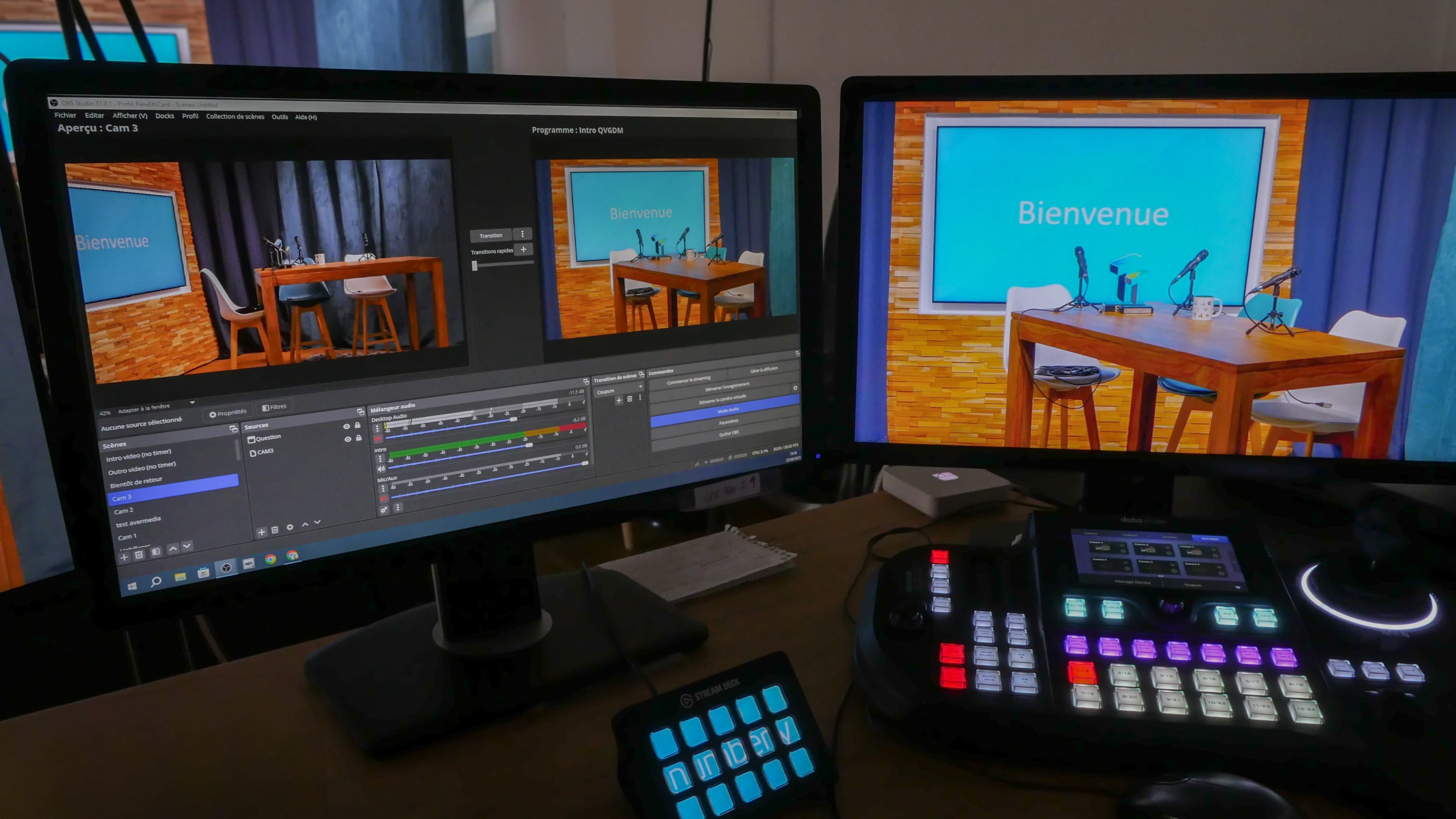 The image shows a dual-monitor setup displaying a video production interface on one screen and a welcome message on a blue background on the other, accompanied by a control panel with various buttons and lights.
